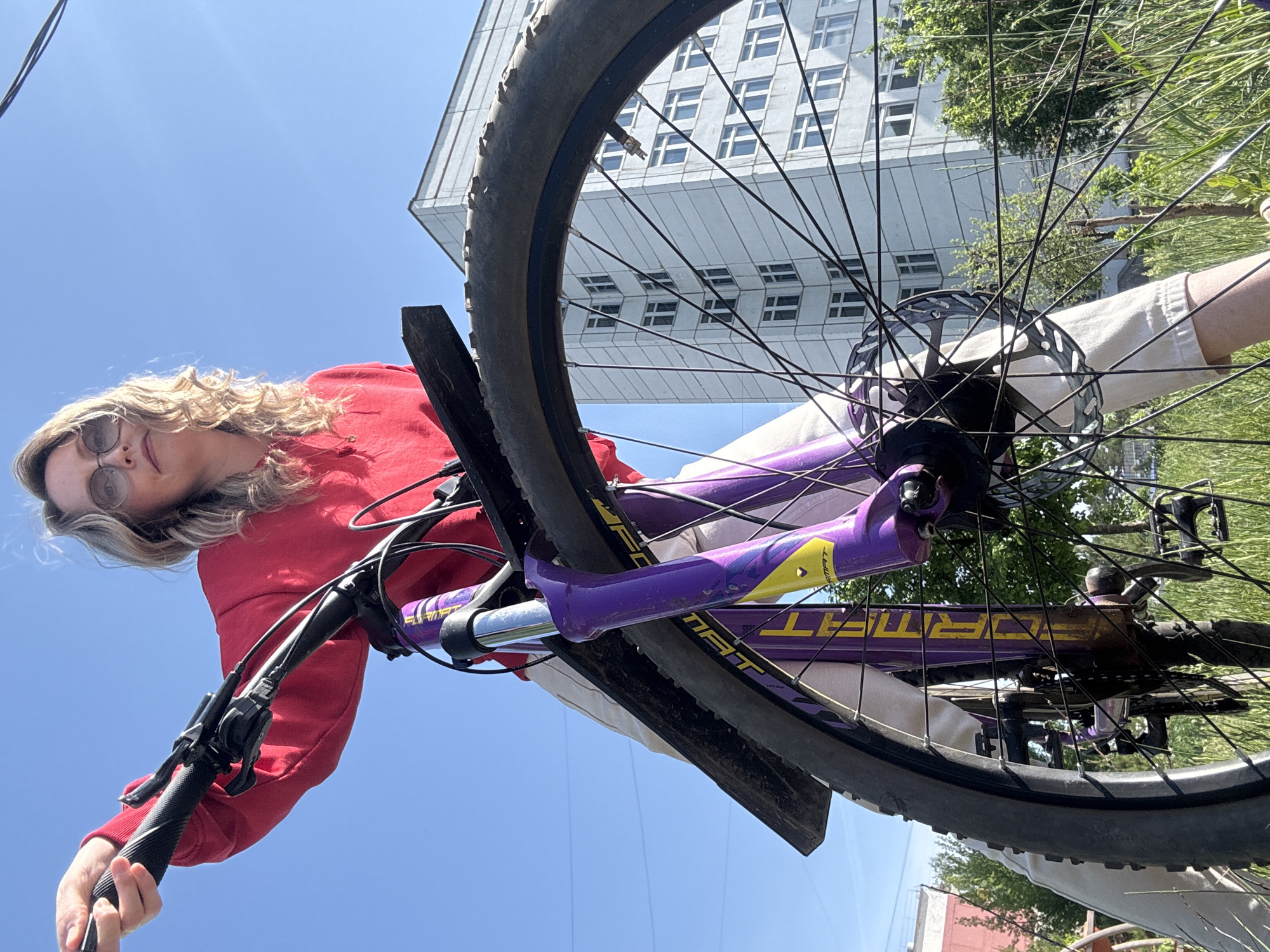 The image presents an upward-angled view of a woman on a bicycle against a backdrop that appears to be in an urban environment, specifically Chaikovsky, Russia.
The woman is fair-skinned, with curly blonde hair and sunglasses. She is wearing a red top and tan-colored pants. She's holding onto the handlebars of the bicycle, suggesting she is either riding or about to ride.
The bicycle is a mountain bike, with a purple frame and yellow accents. The brand "FORMAT" is visible on the frame. The front tire is in clear view, as are the spokes and the braking mechanism.
Behind the bicycle, a tall building with multiple windows is visible. The building is modern in style and suggests an urban area. Some greenery, possibly trees, is visible to the right of the frame.
The sky is blue and appears clear. The photograph is taken during daylight, and the lighting suggests it is a sunny day.