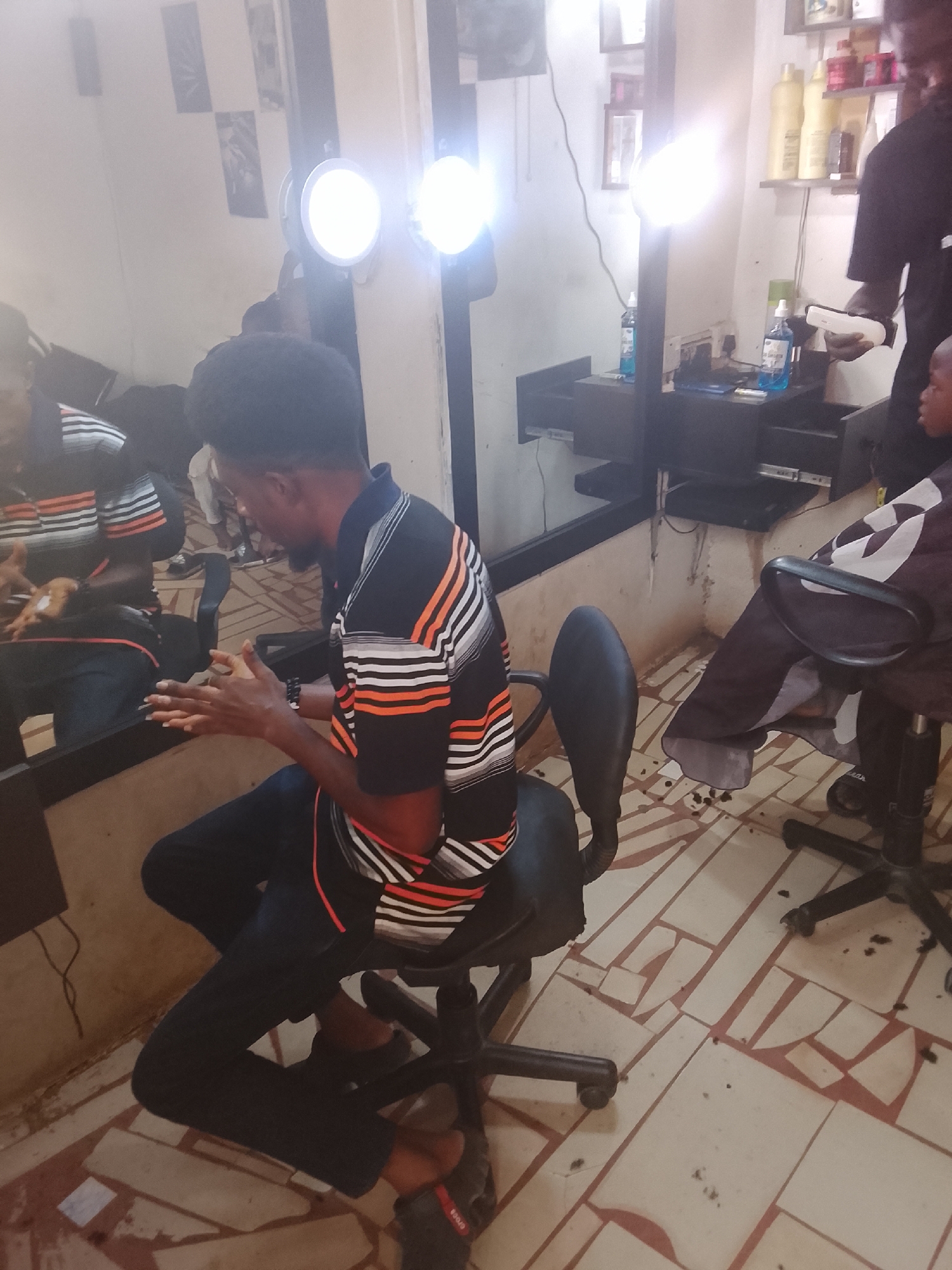 A medium shot depicts the interior of a barber shop in Gwong, Nigeria. A man sits in a barber's chair in the foreground, wearing a black polo shirt with orange and white horizontal stripes. He has dark hair and is facing left, his hands clasped together. A mirror behind him reflects his image.
Behind him and to the right, another man, presumably the barber, is cutting the hair of a young boy seated in another barber's chair. The barber is wearing a black top and the boy is covered with a black and white barber cape.
The shop is equipped with standard barber equipment, including a lighted magnifying lamp, a counter with grooming products, and a mirror spanning the wall. The floor is tiled in a geometric pattern of off-white and reddish-brown. Hair clippings are scattered on the floor around the barber's chairs.