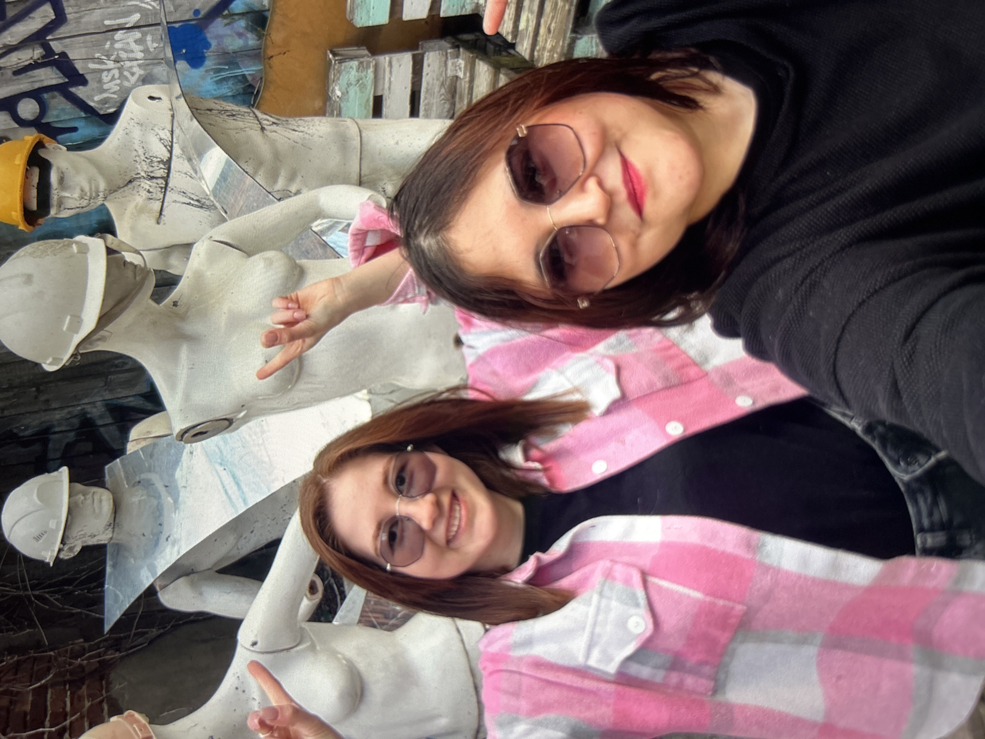 Two young women are taking a selfie in front of several white mannequins in various states of disrepair. The mannequins are wearing hard hats, and one has a yellow hard hat. Some of the mannequins appear to be damaged, with paint splatters and missing parts. The background is a textured wall with graffiti. The woman in the foreground on the left has long brown hair and is wearing a pink and white plaid shirt over a black shirt. She is smiling and wearing sunglasses. The woman on the right has dark brown hair and is also wearing sunglasses. She is wearing a black long-sleeved shirt and is also smiling. The location is described as Unknown, Unknown.