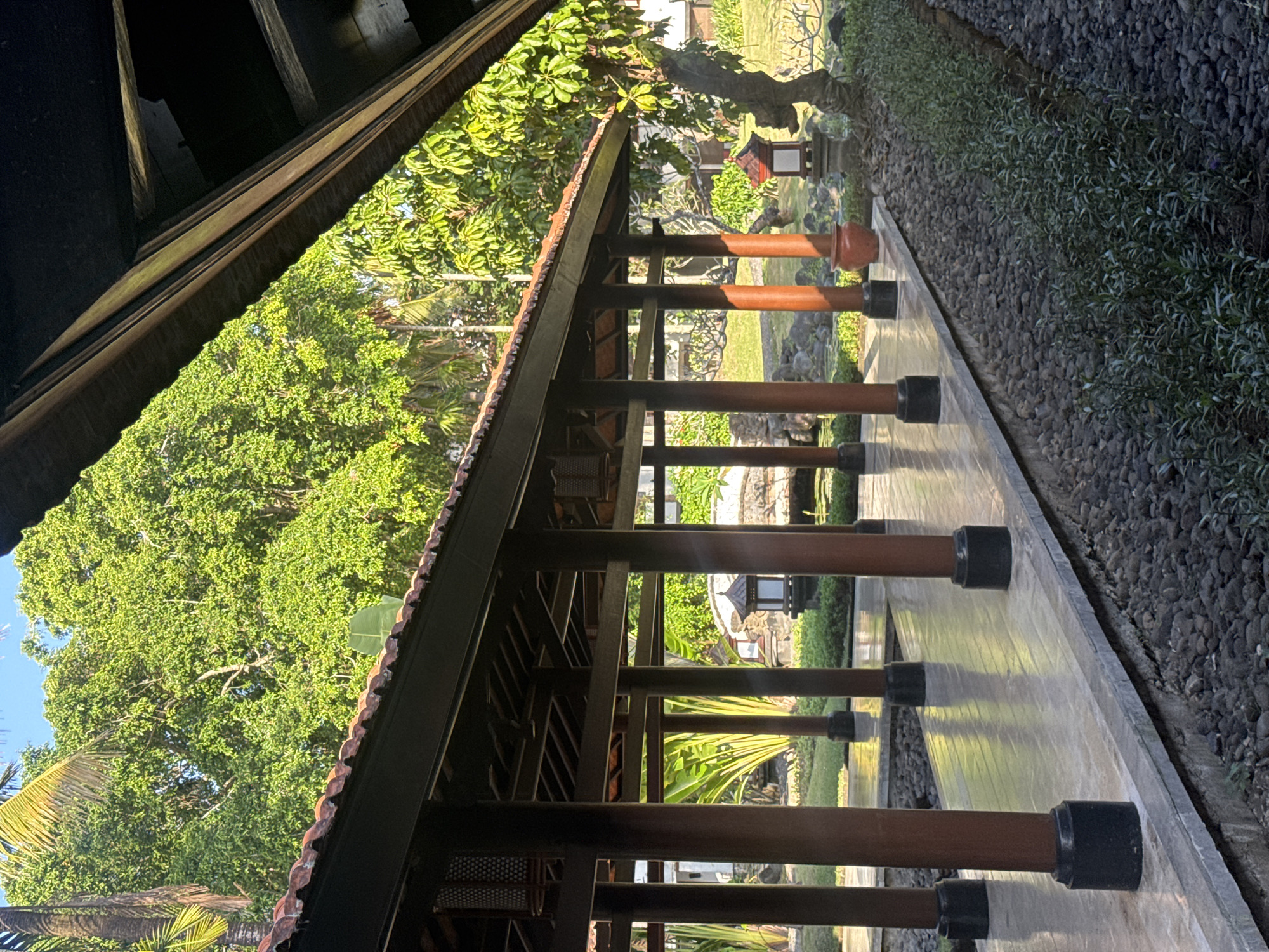 This is a medium shot, eye-level, outdoor photograph of a covered walkway with a tiled roof and dark wooden posts, in Kuta Selatan, Indonesia. The walkway is on the left side of the image and is open to a garden on the right. The ground in front of the walkway is paved with smooth, light-colored tiles that reflect the sunlight. To the right of the tiled walkway, there is a border of dark grey pebbles, and beyond that, a row of small, green plants. Further into the garden, there are large trees with lush green foliage, a stone pathway, and decorative stone lanterns. A large, terracotta pot is visible near the walkway. The sky is bright blue with a few wisps of clouds. The overall scene suggests a resort or a tranquil garden setting.
