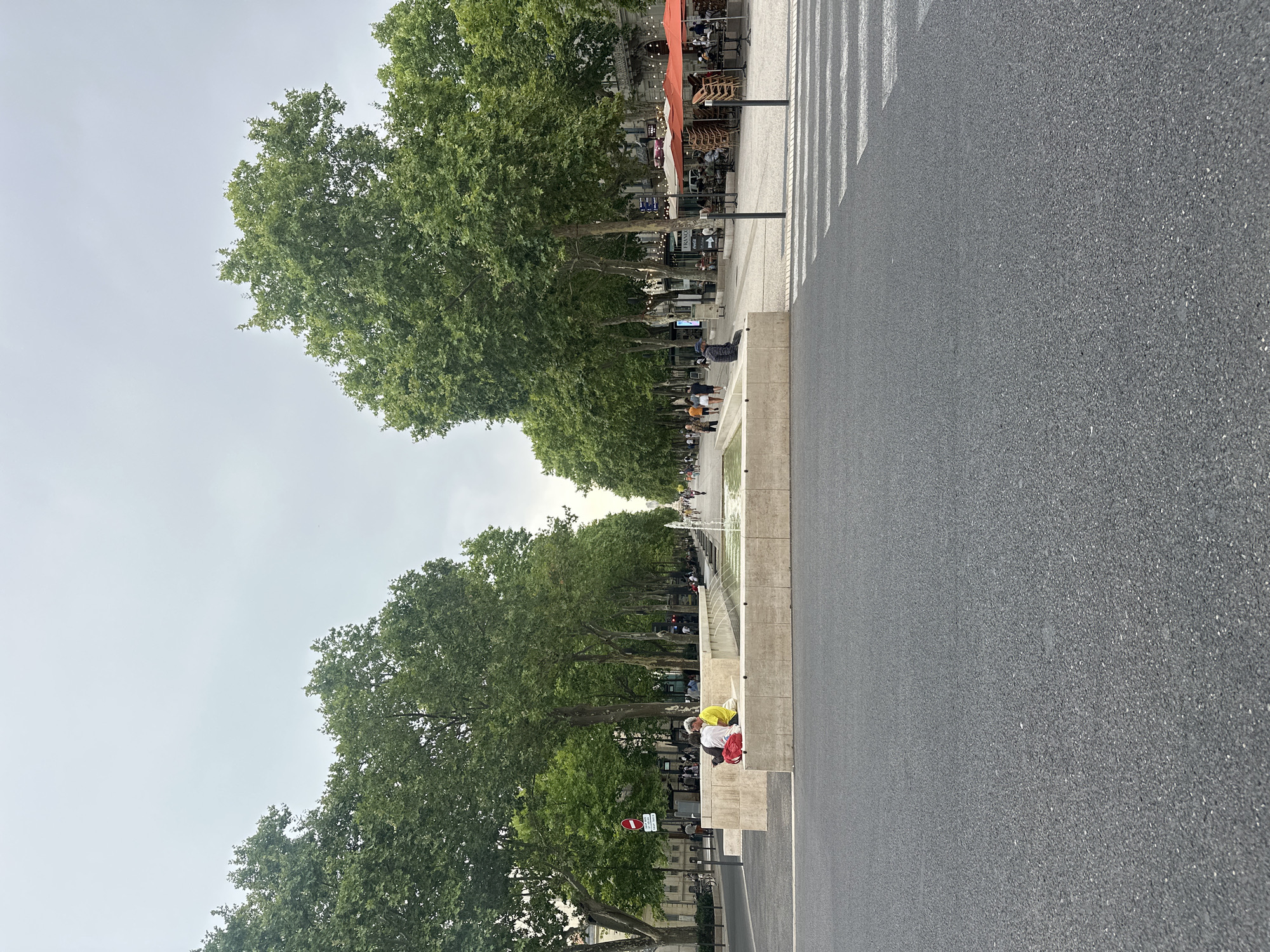 The image depicts a wide, tree-lined avenue in Nîmes, France, likely Place de la Maison Carrée, on an overcast day. In the foreground, a section of asphalt road with a pedestrian crossing is visible. Beyond the road, a long, low, L-shaped stone planter with a fountain running through it separates the pedestrian walkway from the road. Two individuals are seated on the planter, one wearing a yellow shirt and the other in a dark shirt. In the background, the avenue is flanked by mature trees casting shadows on the paved walkway. Numerous people are strolling along the avenue, and several outdoor cafe tables with orange umbrellas are set up on the right side, suggesting a commercial area. The architecture visible on the sides of the avenue appears to be of historical significance, consistent with the city's Roman heritage.