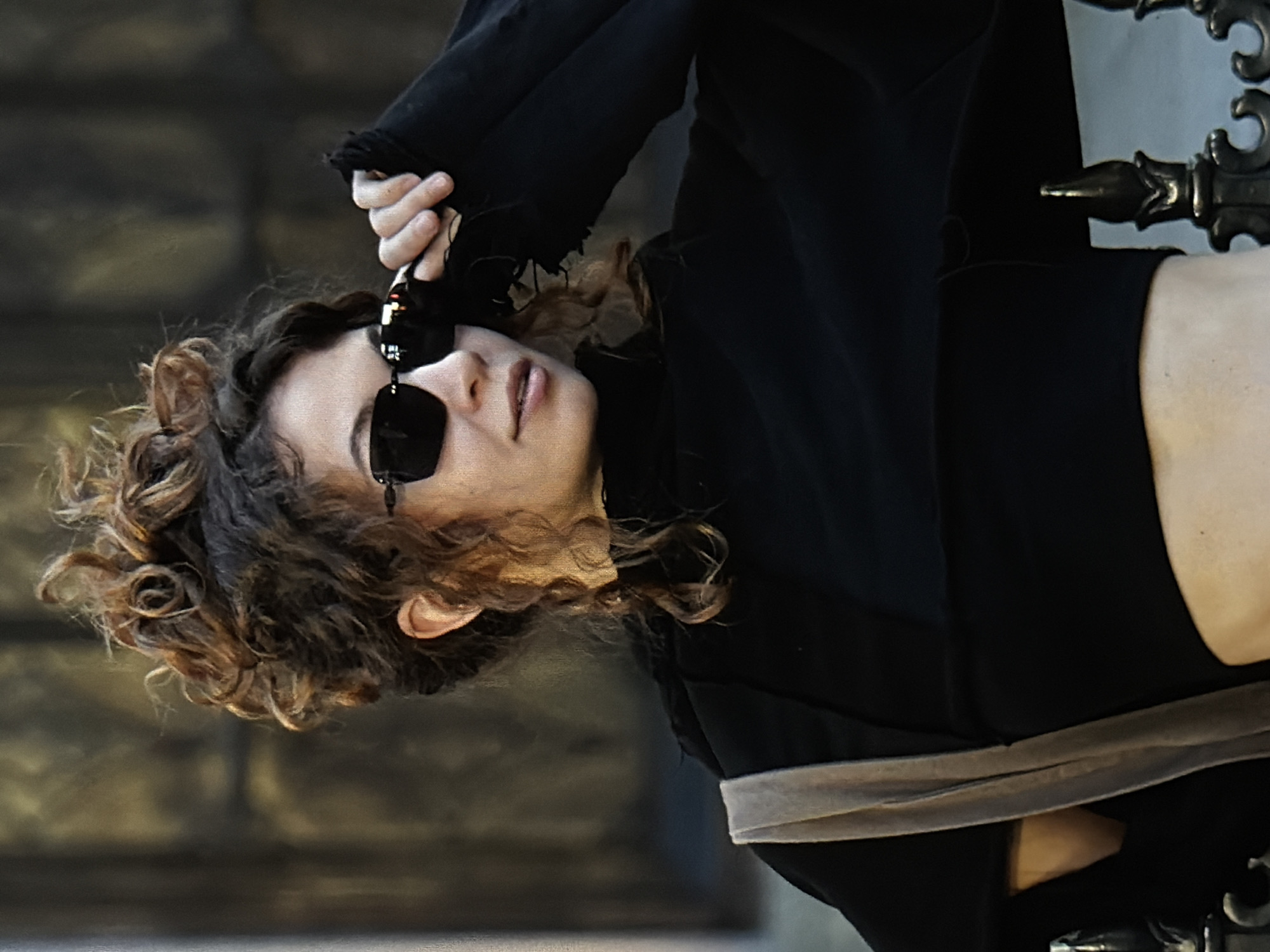 A woman with curly brown hair and wearing black sunglasses is holding the sunglasses by the frame in her right hand. She is dressed in a black top and appears to be sitting on a decorative metal chair or bench. The background is a textured, dark-colored wall, possibly stone or concrete. The location is not discernible as Đà Nẵng, Vietnam, from this image alone.