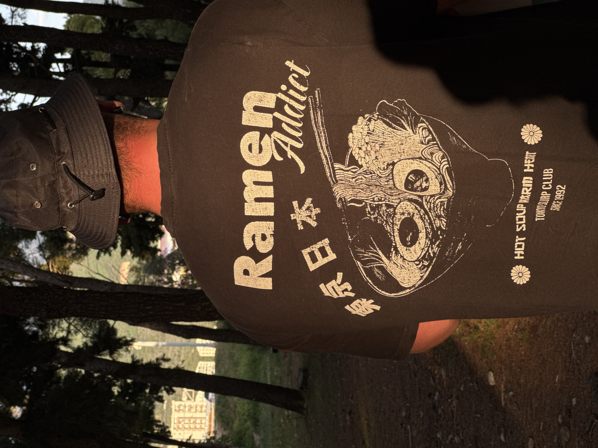 The image, taken in Bar, Montenegro, shows a person from behind, wearing a dark-colored T-shirt and a dark bucket hat. The person's back, neck, and the side of their head are visible. The setting is outdoors, likely an elevated area or a park, characterized by numerous dark-trunked trees that frame the view. In the distant background, several multi-story buildings are faintly discernible, indicating an urban or suburban environment.
The time of day appears to be late afternoon or early evening, as suggested by the warm, golden light illuminating parts of the scene, particularly the person's right side and neck. The weather seems clear and bright.
The most prominent detail is the graphic design on the T-shirt. Large white text arranged vertically reads "Ramen Addict." Below the word "Ramen," Japanese characters "日本製" (Nihon sei), meaning "Made in Japan," are displayed. To the right of the text is a stylized illustration of a ramen bowl, featuring noodles forming two large, round, eye-like shapes, along with other ramen ingredients such as seaweed and what appear to be sprouts, and chopsticks. Further to the right, smaller white text states "HOT SOUP WARM HEAT," followed by "TOKYO SOUP CLUB," and "SINCE 1992." A small, stylized flower or star emblem is positioned next to "HOT SOUP WARM HEAT." The person's reddish-brown hair is visible on the back of their neck beneath the hat. The overall scene suggests a casual outdoor moment.