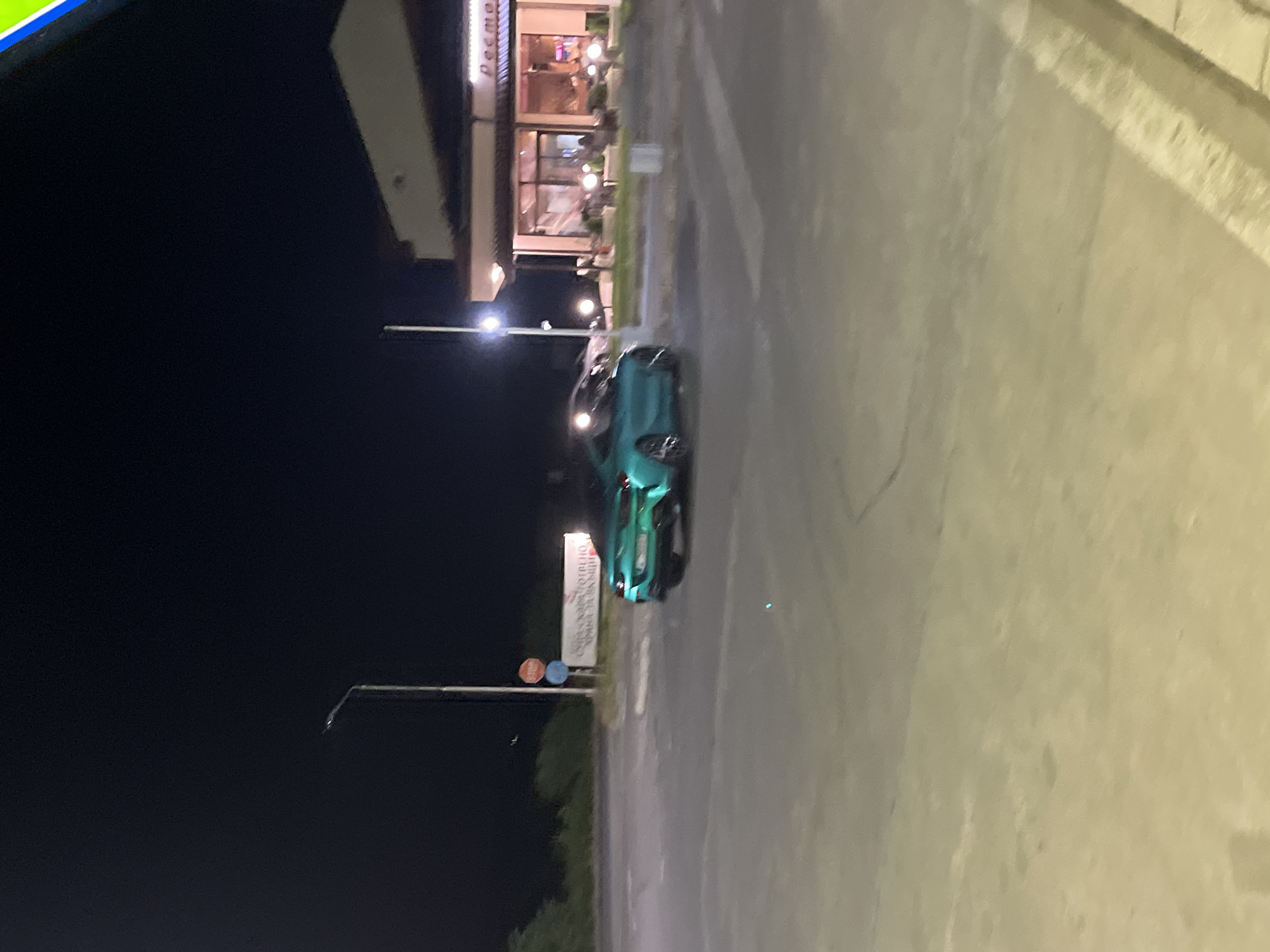 The image shows a nighttime scene in Popovitsa, Bulgaria. A light-green car is parked on the side of a road next to a sign. The road has a concrete curb on one side. In the background, there is a building with outdoor lights and a sign that appears to read "Peemo." A street lamp is visible. The sky is dark.