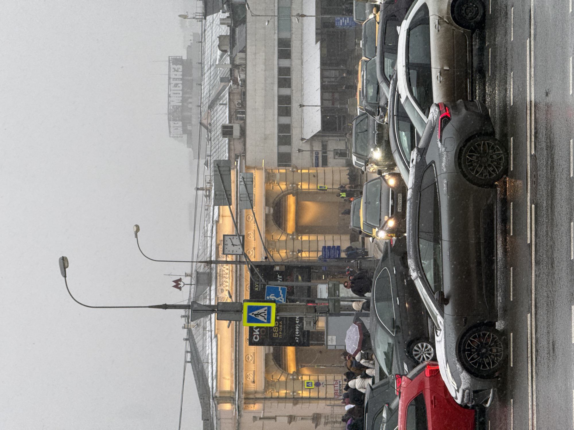 The image depicts a street scene in Moscow, Russia, during what appears to be snowy weather. The focus is on a line of cars stuck in traffic on a wet road. The architectural style of the buildings visible suggests a Russian cityscape.
On the left side of the image, a large, illuminated building with archways can be seen. This is likely a historical or significant structure, possibly a train station or government building. Streetlights are present, with a metro sign. Pedestrian crossing signs are also visible.
People are seen walking and standing on the sidewalks, some holding umbrellas. Various shops and advertisements are visible. The building in the background has the word "MOSTAZ" in Cyrillic, suggesting it is some kind of building.