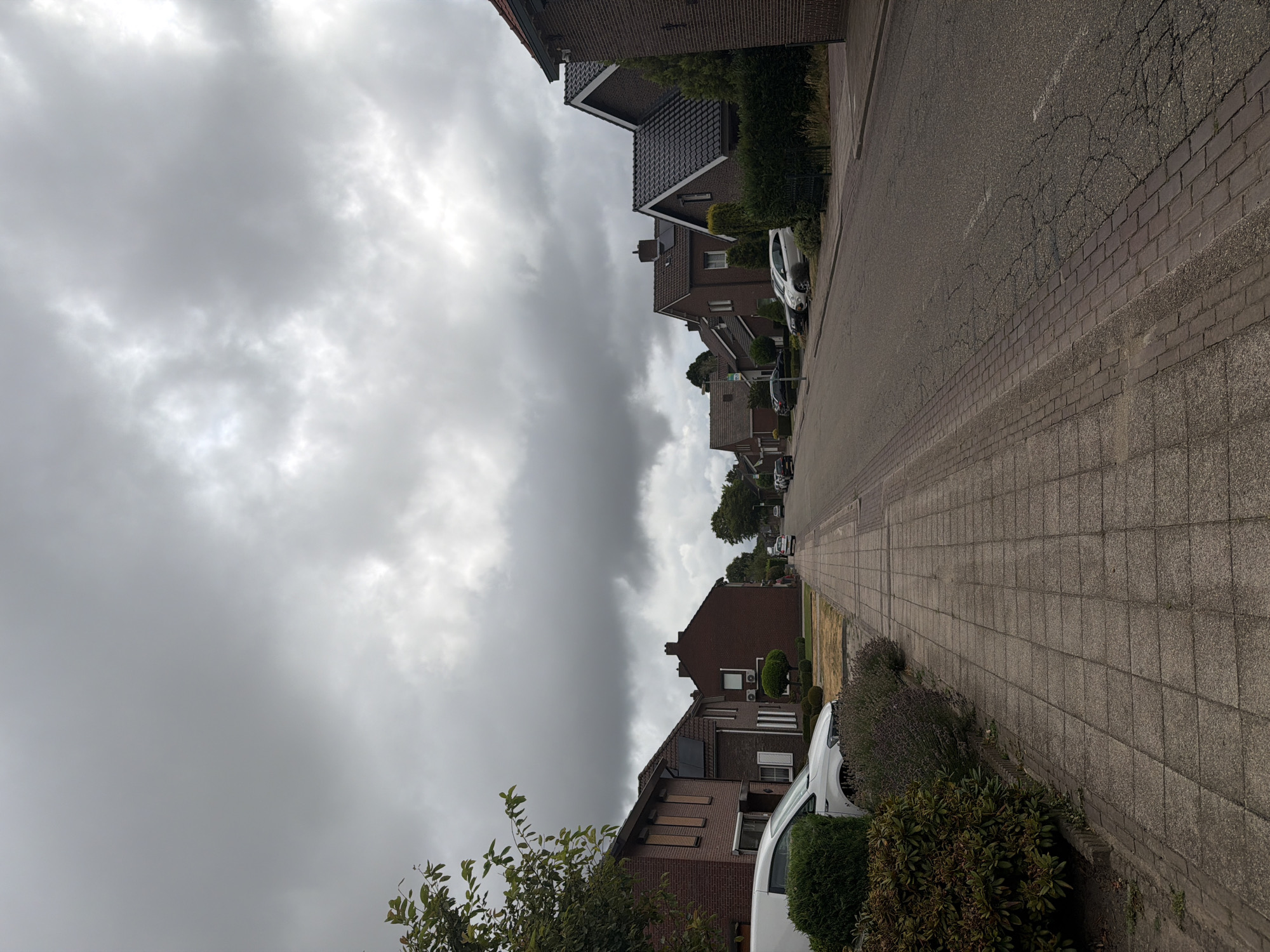 The image depicts a residential street in Melick, Netherlands, under a heavily overcast sky. A paved sidewalk runs along the left side of the street, bordered by manicured hedges and bushes. Several houses with red brick exteriors and dark tiled roofs are visible lining the street. Parked cars are present on both sides of the road. The street itself is asphalt, showing signs of wear with visible cracks. Trees and foliage are scattered throughout the scene, adding greenery to the urban landscape. The overall impression is one of a quiet, suburban neighborhood on a cloudy day.
