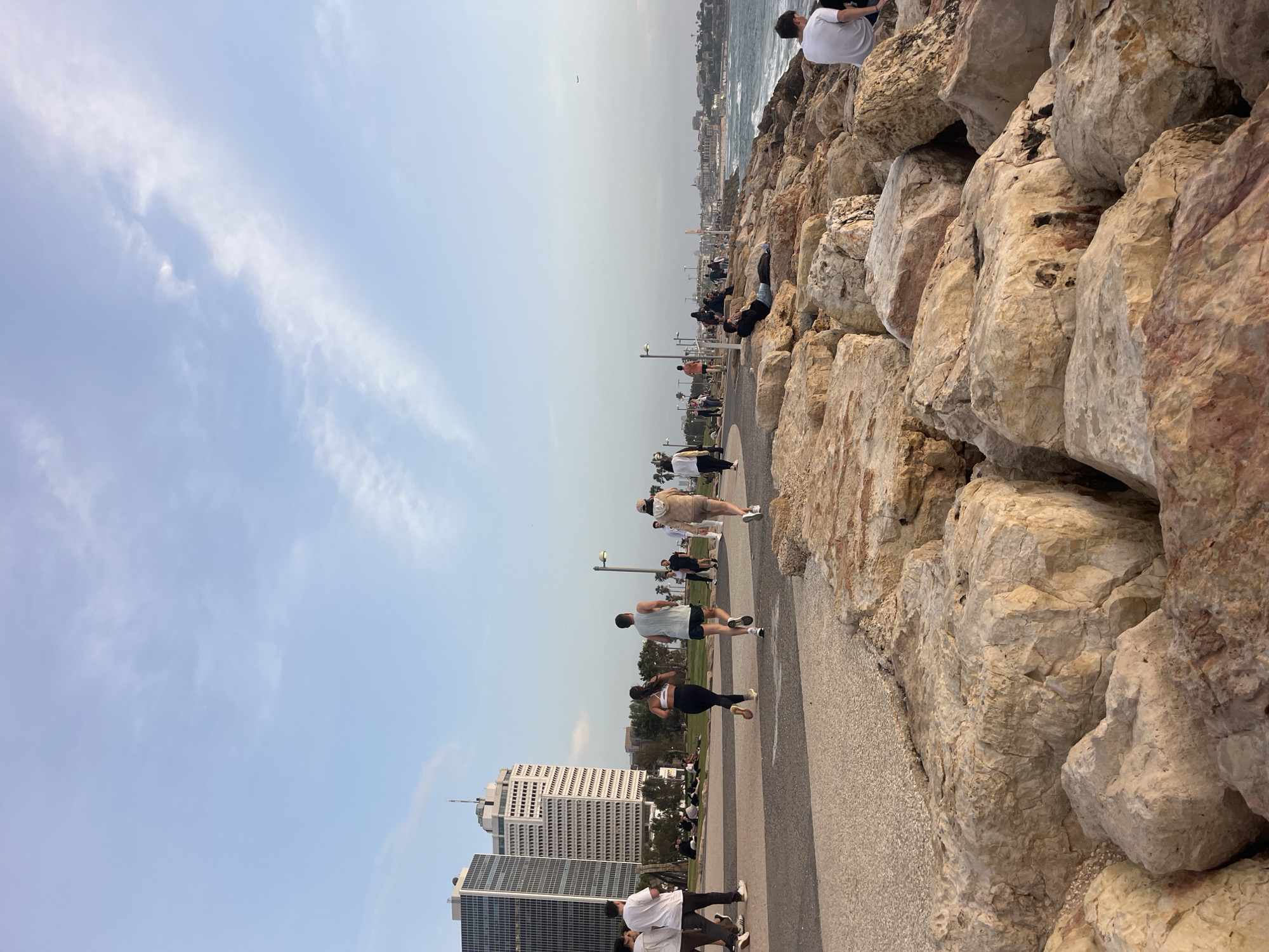 The image depicts a coastal scene in Tel Aviv, Israel. A wide paved pathway runs along the right side, bordered by large, light-colored rocks that separate it from the sea. People are walking, jogging, and sitting along the pathway. Several tall buildings are visible in the background towards the left side of the image. The sky is light blue with scattered clouds.
