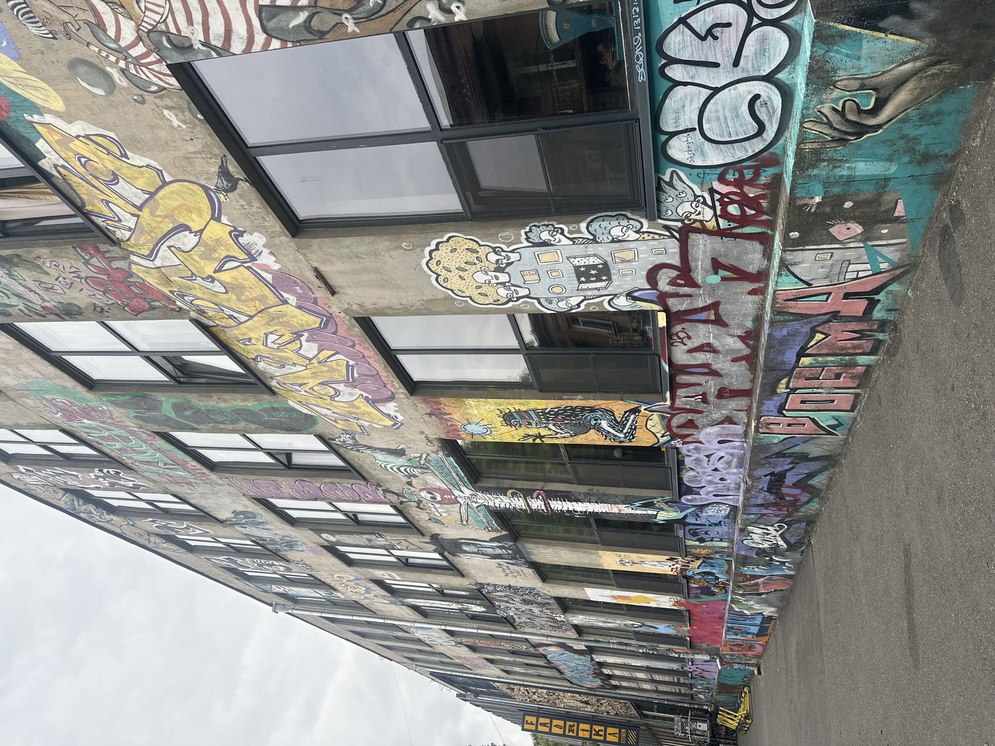 The image captures a multi-story building facade in Tbilisi, Georgia, heavily adorned with vibrant graffiti art. The building itself appears to be an older, industrial-style structure with a concrete exterior and numerous large, dark-framed windows. The graffiti covers almost every visible surface, from the lower concrete barrier wall running along the sidewalk to the upper levels of the building.
The art styles vary widely, including bold, bubble-style lettering in colors like yellow, purple, and pink, as well as more intricate figurative drawings and abstract designs. Among the graffiti, recognizable elements include a zebra, a frog, a figure with a large afro and multiple faces, and a depiction of hands. The building is situated along a paved sidewalk or street, with a small portion of yellow electric scooters visible in the background to the left. A vertical sign for "FABRIKA HOSTEL" is also visible on the left side of the building, indicating the location. The sky is overcast, suggesting a cloudy day. No people are clearly discernible or actively engaged in any observable actions within the frame. The overall scene conveys a sense of urban decay and artistic expression.