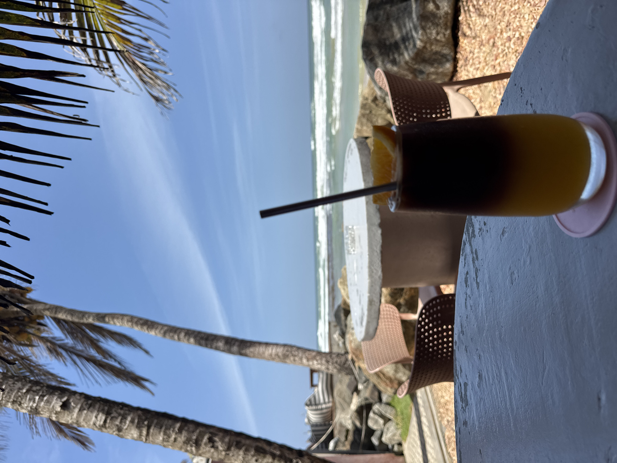 The image captures a serene beachside setting in Ahangama, Sri Lanka. In the foreground, a tall glass of layered beverage, likely a coffee concoction with orange juice at the bottom and dark coffee on top, is garnished with an orange slice and a straw. It sits on a pink coaster placed on a weathered, light-colored table. Behind this table, a round, light-colored concrete table is visible, accompanied by two chairs, one in a muted pink and the other a darker brown. The background reveals the ocean with gentle waves breaking on the shore, under a bright blue sky dotted with thin clouds. Two palm trees with textured trunks and fronds frame the left side of the scene, with their leaves extending towards the top of the image. Rocks and patches of green grass are scattered along the beach. The overall ambiance is one of a relaxed, tropical cafe by the sea.