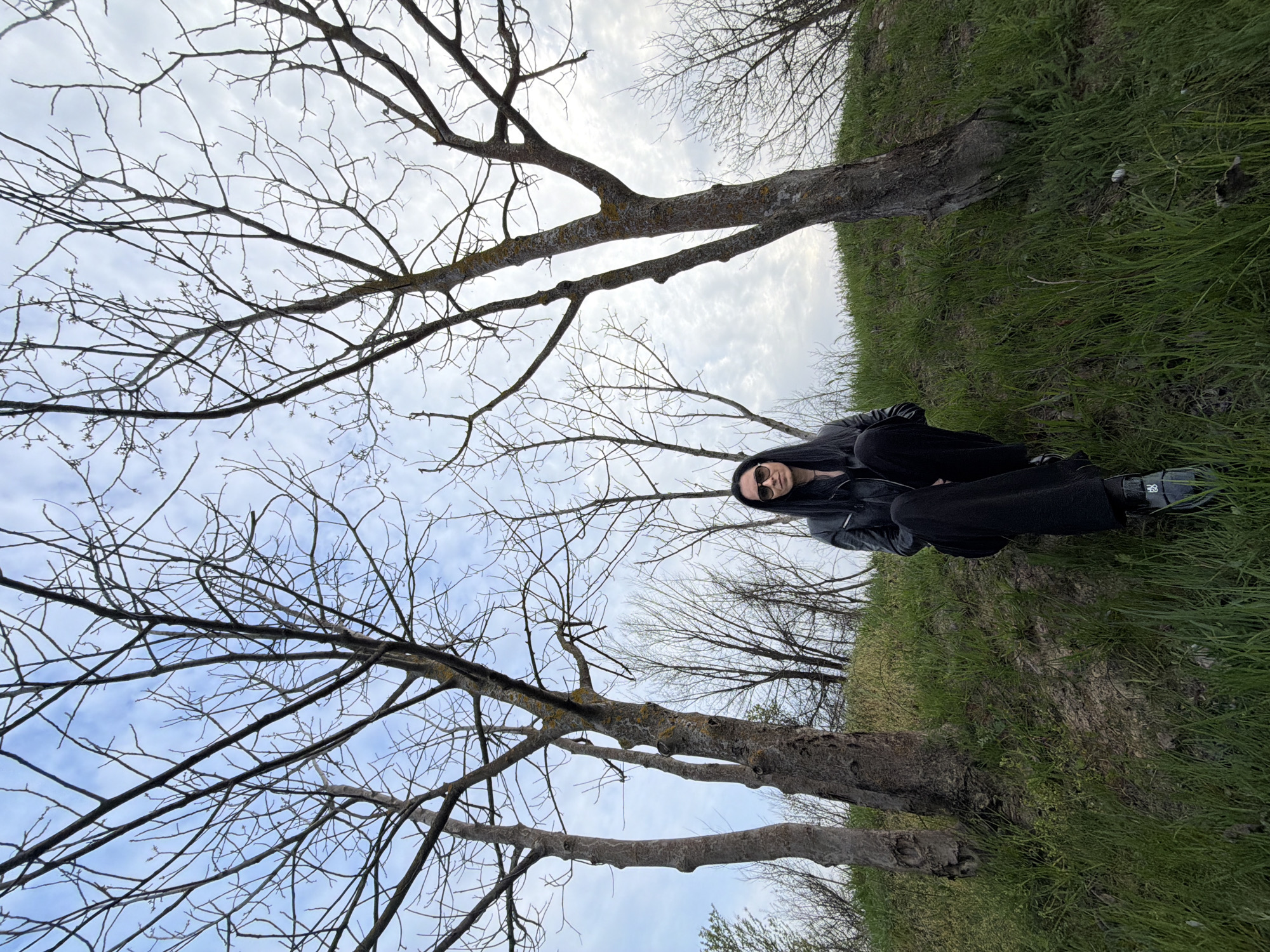 A person wearing black clothing and sunglasses is sitting on a grassy area between two bare trees. The sky is visible through the branches of the trees, and the overall environment appears to be outdoors in a park or wooded area. No specific details indicating the location is in Pleven, Bulgaria, are readily apparent in the image.
