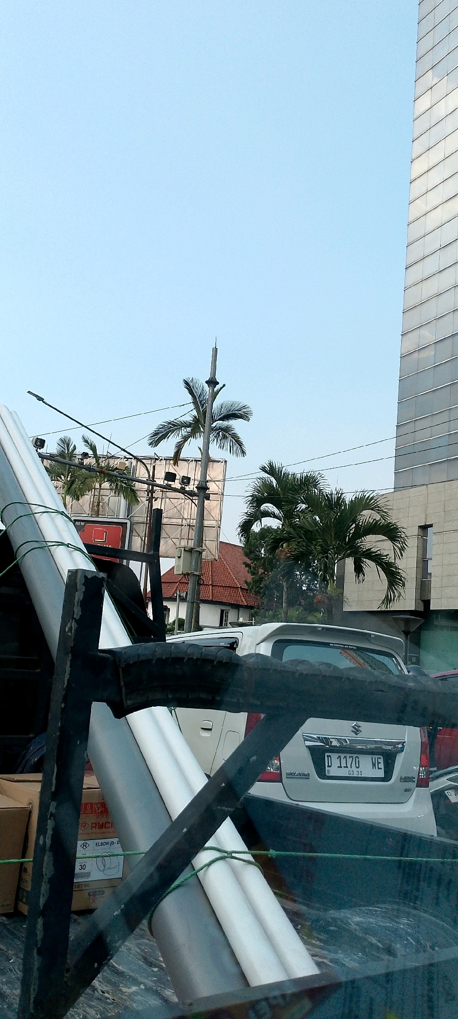 The image captures a street scene in Bandung City, Indonesia, under a clear blue sky. In the foreground, a dark metal frame obscures part of the view. Beyond the frame, a truck bed contains several large, cylindrical objects, one gray and two white, secured with green rope. To the right of the truck bed, a white Suzuki car with a license plate reading "D 1170 WE" is visible. In the background, palm trees stand prominently, with a large, modern glass-building to the far right. A billboard structure is also visible behind the palm trees. The overall environment suggests an urban setting with a mix of natural elements and modern architecture.