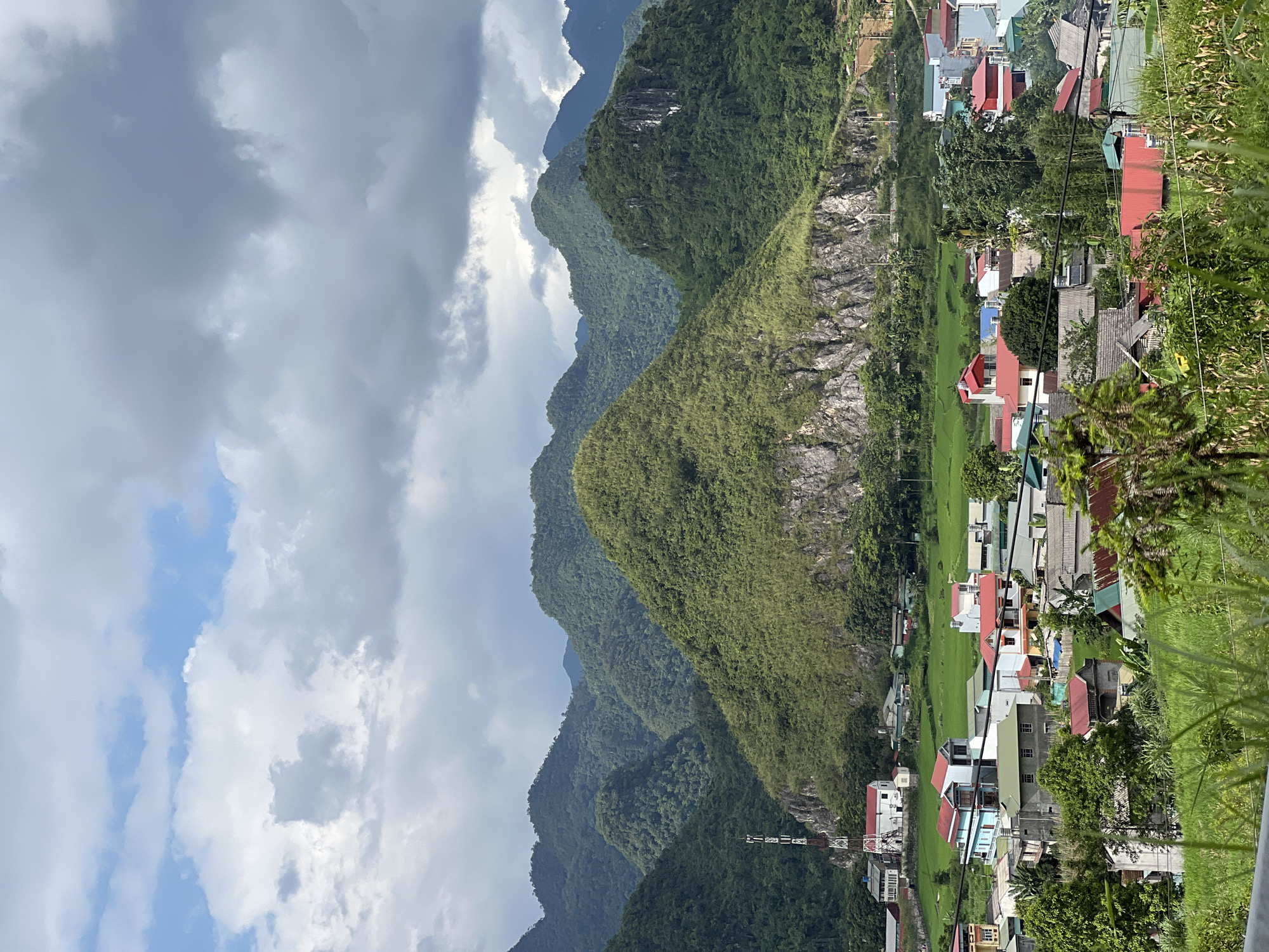 The image depicts a panoramic view of a rural village nestled in the Quản Bạ Commune of Vietnam, against a backdrop of lush, green, undulating mountains. The sky is partly cloudy, with dramatic formations of grey and white clouds dominating the upper portion of the frame. In the mid-ground, a prominent, conical-shaped mountain covered in dense vegetation rises sharply, its rocky base exposed in places. A communication tower stands to the left of this central mountain.
Below the mountains, the village is characterized by a cluster of houses with varying architectural styles and vibrant roof colors, predominantly red and blue, interspersed with green foliage. The houses appear to be constructed from concrete and wood. Surrounding the village are rectangular fields of green, likely rice paddies, indicating agricultural activity. The foreground features the tops of green plants and some corn stalks, suggesting the view is from a slightly elevated position overlooking the scene. The overall impression is one of a serene, rural landscape with the distinct geological formations of the region.