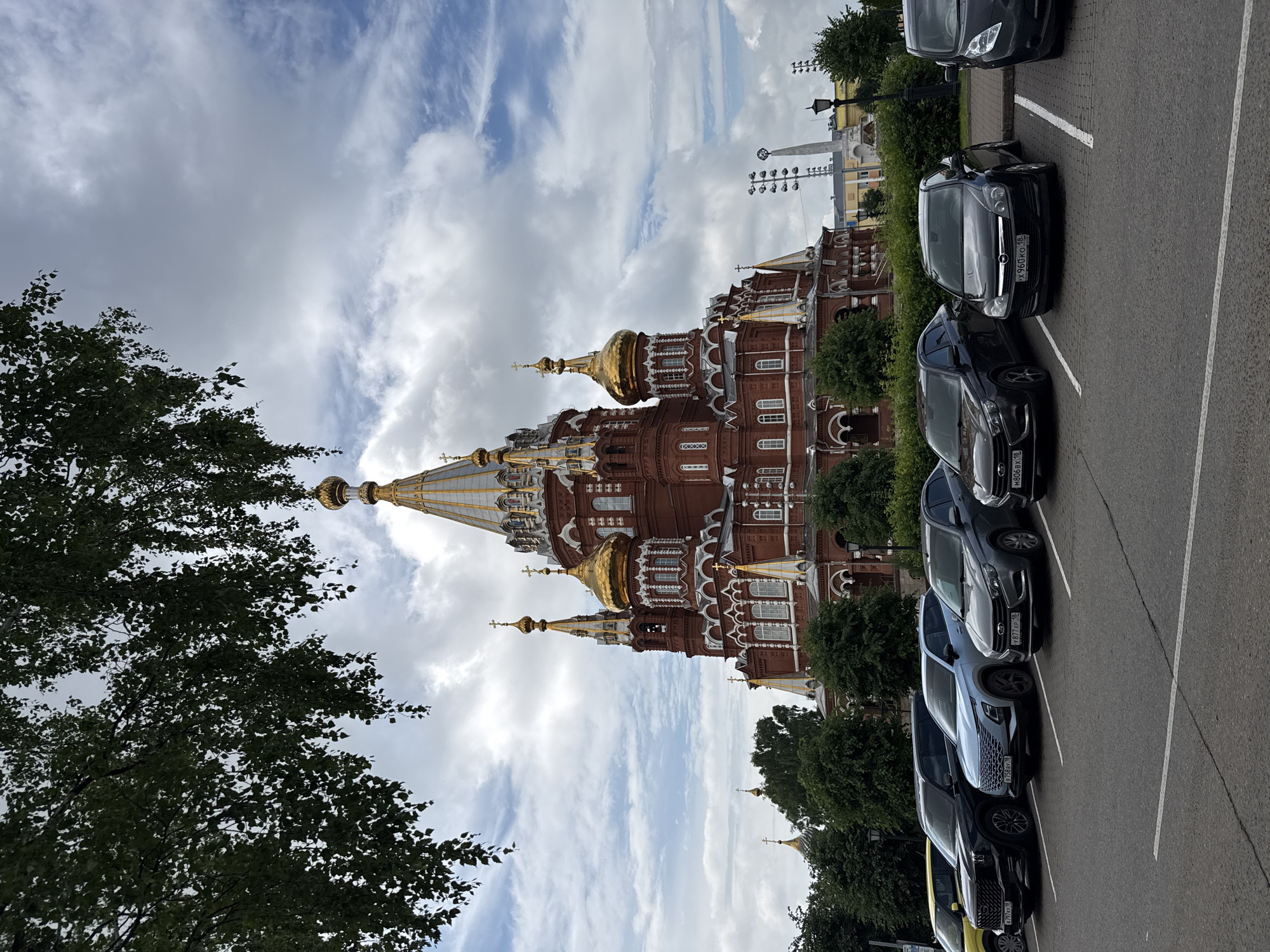 The image captures the Alexander Nevsky Cathedral in Izhevsk, Russia, a prominent red-brick Orthodox church adorned with golden domes and white decorative elements. In the foreground, a row of parked cars, including a yellow sedan, a black SUV, a light blue SUV, and several dark sedans, are visible in a parking lot. The sky above is partly cloudy, with white clouds scattered across a blue expanse. Lush green trees frame the scene, with some branches extending into the upper left corner of the image. In the background, beyond the church, a tall structure with lights and a monument can be seen, along with street lamps and more trees. The overall impression is of a clear day, likely during daylight hours, with the cathedral serving as the central focal point.