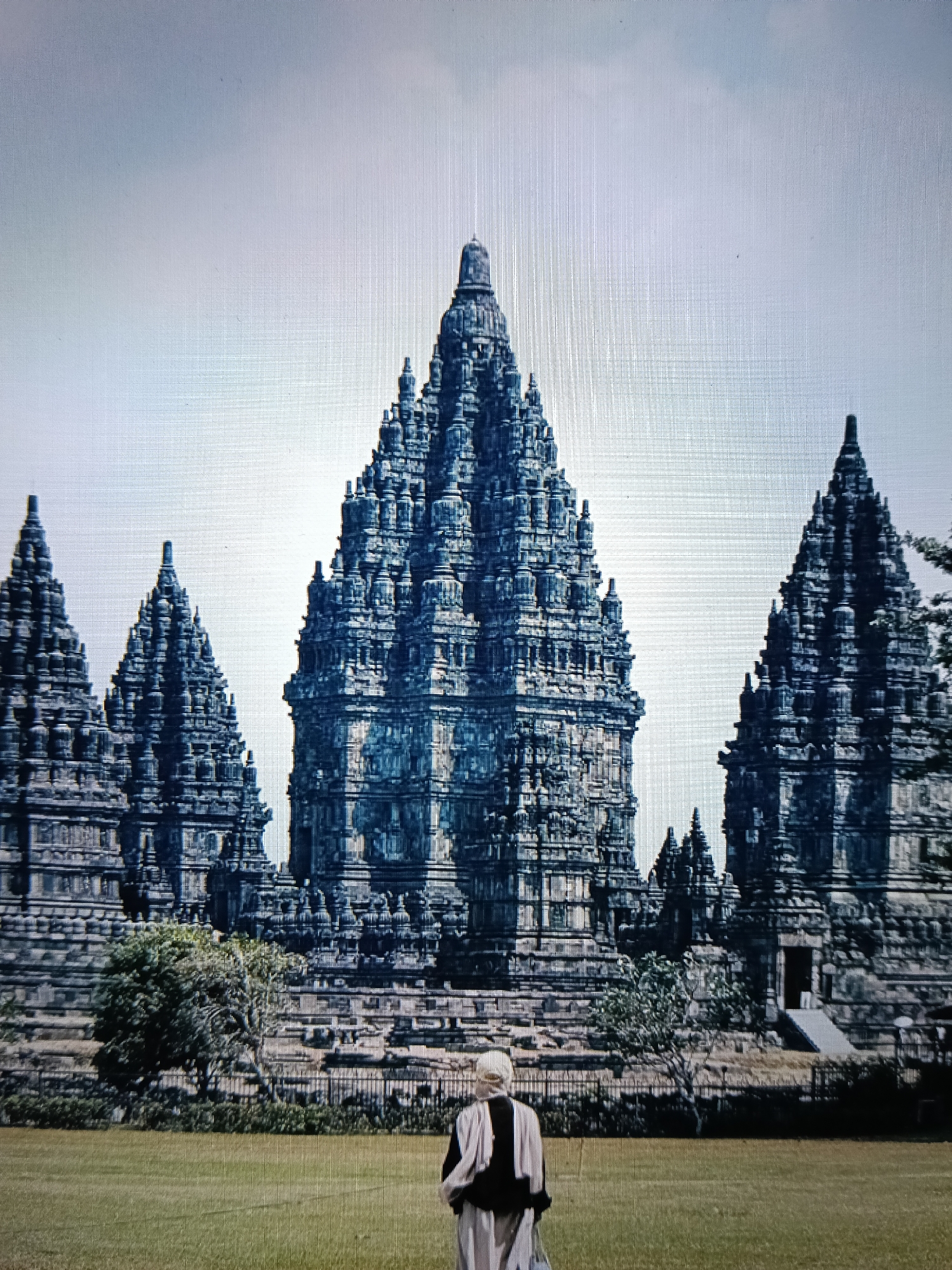 The image depicts a serene outdoor scene featuring the ancient Prambanan Temple complex, a Hindu temple compound located in Yogyakarta, Indonesia, and not in Raha. The temple, characterized by its towering, intricately carved spires, is a dominant feature in the background. The architecture is distinctly Javanese, with numerous smaller temples surrounding the central, largest structure. The temple complex appears to be constructed from dark stone, with visible detailing and ornamentation.
In the foreground, a person is standing with their back to the camera, facing the temple. The individual is wearing a light-colored garment, possibly a robe or a long tunic, with dark accents around the shoulders and collar. Their hair is visible at the back of their head, appearing light in color. The person is positioned on a vast expanse of well-maintained green grass, suggesting a park-like setting.
The environment around the temple includes some sparse vegetation, including trees and shrubs, with a fence line separating the grassy area from the temple grounds. The sky is overcast with a light, diffused light, indicating a cloudy or hazy day. The image exhibits a slightly pixelated quality, hinting at its capture through a digital medium or a projection. The overall impression is one of historical grandeur and quiet contemplation.