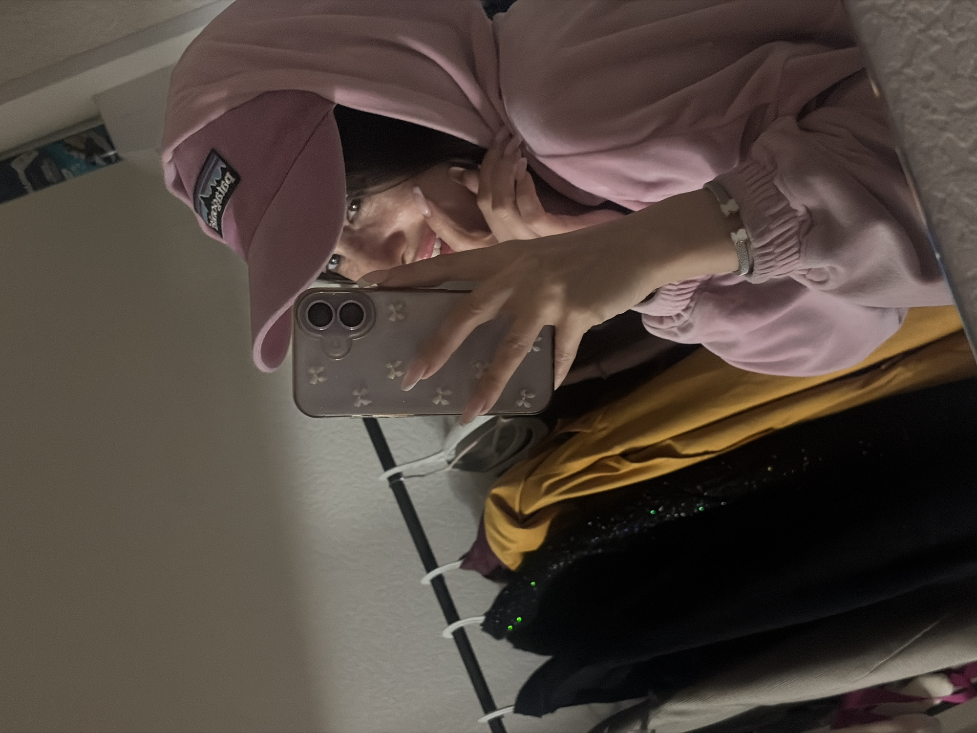 A mirror selfie shows a person wearing a pink hooded sweatshirt and a light pink baseball cap with a dark logo on the left side. The person is holding up a smartphone with a light-colored case. Their face is visible, with dark hair peeking out from under the hat. A silver bracelet is on their left wrist. Behind the person, there is a clothing rack with several hanging garments, including items in yellow, black, and gray tones. The location of the city is "Unknown, Unknown". The environment appears to be inside a room with a light-colored wall.