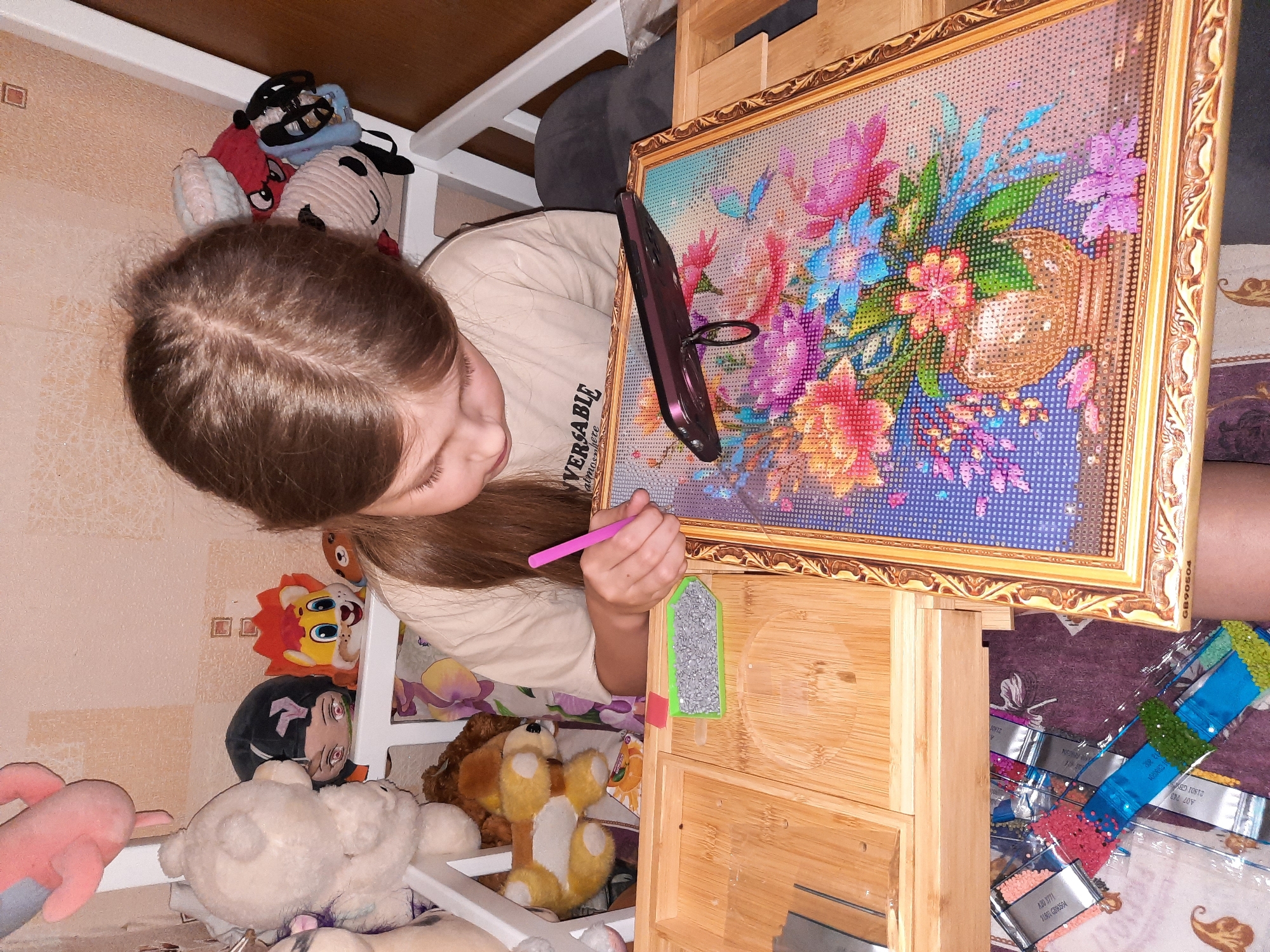 A young girl with long brown hair is engaged in a diamond painting activity. The painting, framed in ornate gold, displays a colorful floral arrangement. She holds a pink tool in her right hand, hovering over the painting. Her cell phone is mounted on the upper right of the painting. Next to the painting is a small wooden organizer, holding a tray of diamond beads. Diamond painting supplies are stacked around the base of the organizer. The scene is situated on a bed or platform surrounded by stuffed animals. The room appears to be indoors, possibly a bedroom. The image location is the city of Izhevsk, Russia.