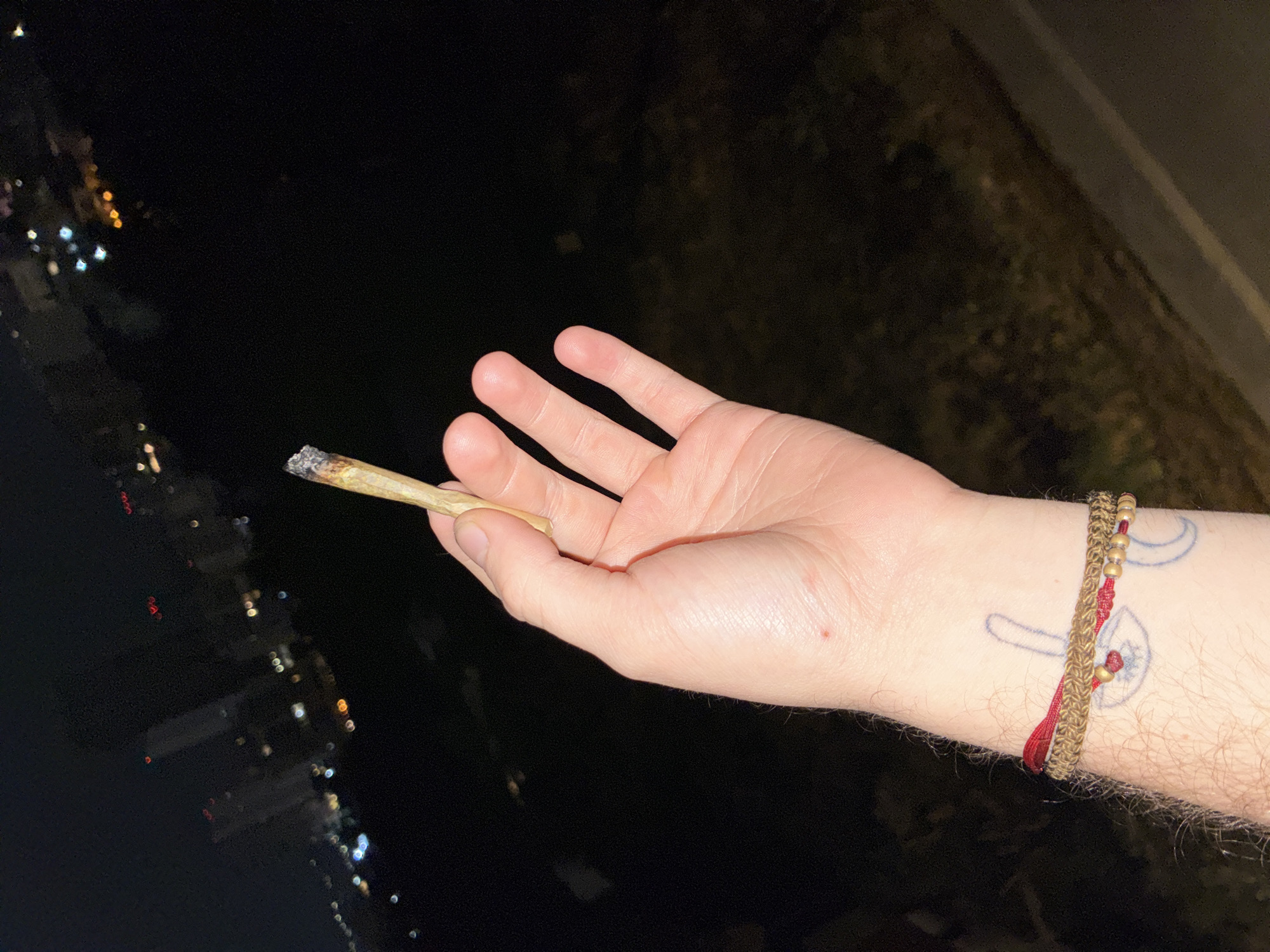 A person is holding a lit marijuana cigarette in their left hand, with a city skyline in the background, likely at night. The person has a tattoo and a bracelet on their left wrist. The location is probably in Huixquilucan de Degollado, Mexico.