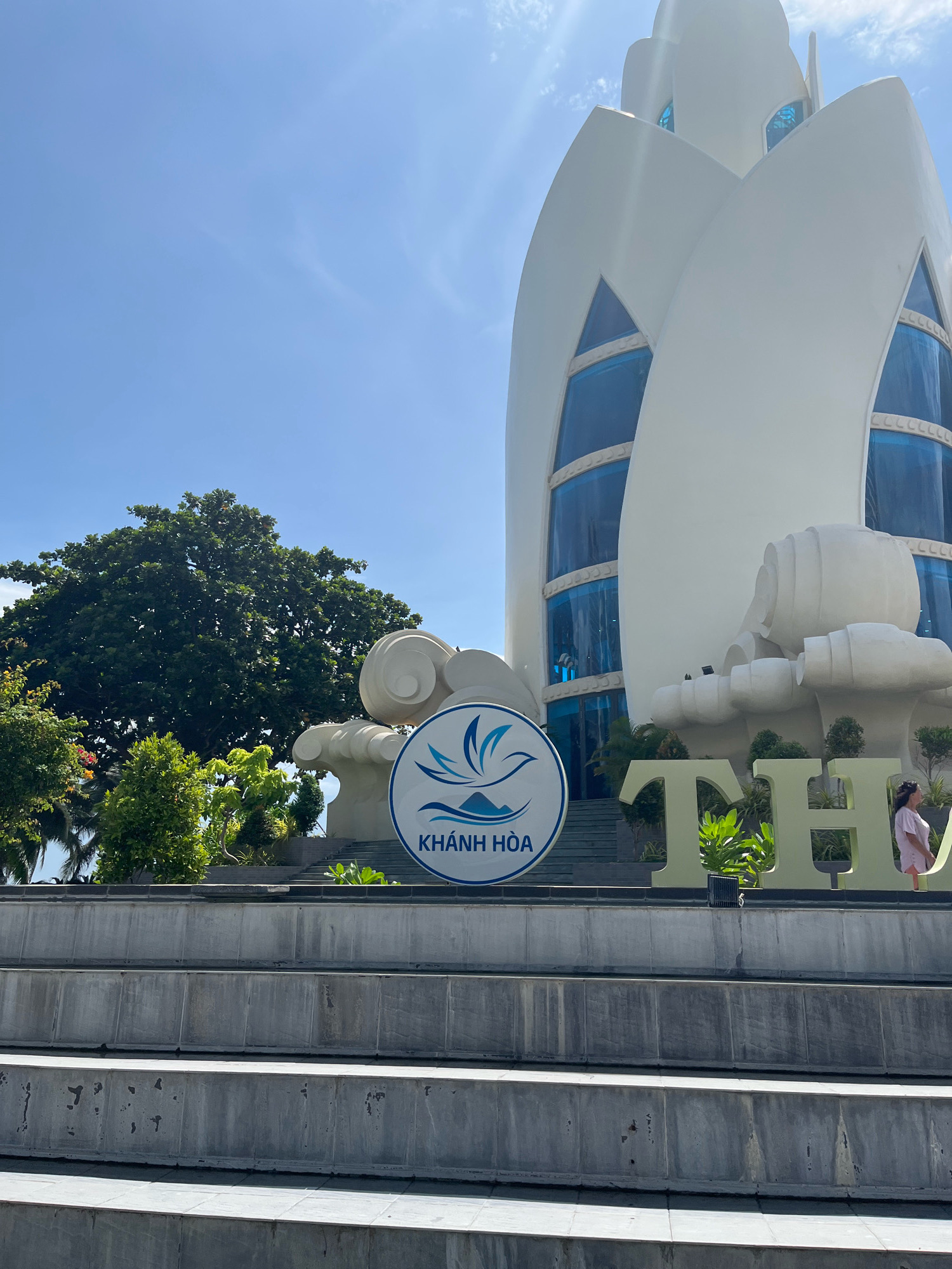 The image depicts the entrance to the Nha Trang Center, a prominent landmark in Nha Trang, Vietnam. The building, with its distinctive white, petal-like architectural design and blue-tinted windows, stands tall against a clear blue sky. In the foreground, a circular sign with the logo for "Khánh Hòa" is prominently displayed, featuring a stylized bird in flight over mountains and waves. Large, gold-colored letters spelling "THAI" are also visible to the right of the sign, suggesting a connection to a resort or hotel. Several steps lead up to the building, and lush green trees and other vegetation surround the area. A woman in a pink outfit is visible on the right side of the image, looking towards the building. The overall scene conveys a sense of arrival and attraction, highlighting the unique architecture and local branding of Nha Trang.