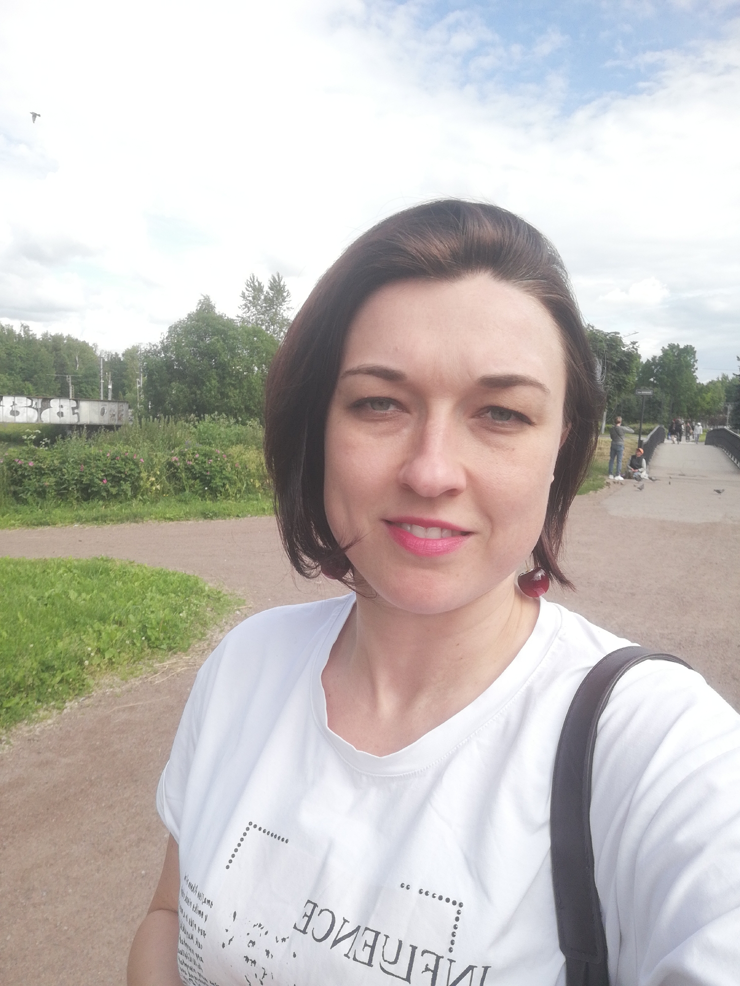 A woman with dark brown hair and blue eyes, wearing a white t-shirt with black text and cherry-shaped earrings, stands on a dirt path in a park. In the background, there is lush green vegetation, trees, a bridge, and people walking. The sky is cloudy with patches of blue. The overall scene depicts a casual outdoor setting in a park, possibly in a city.