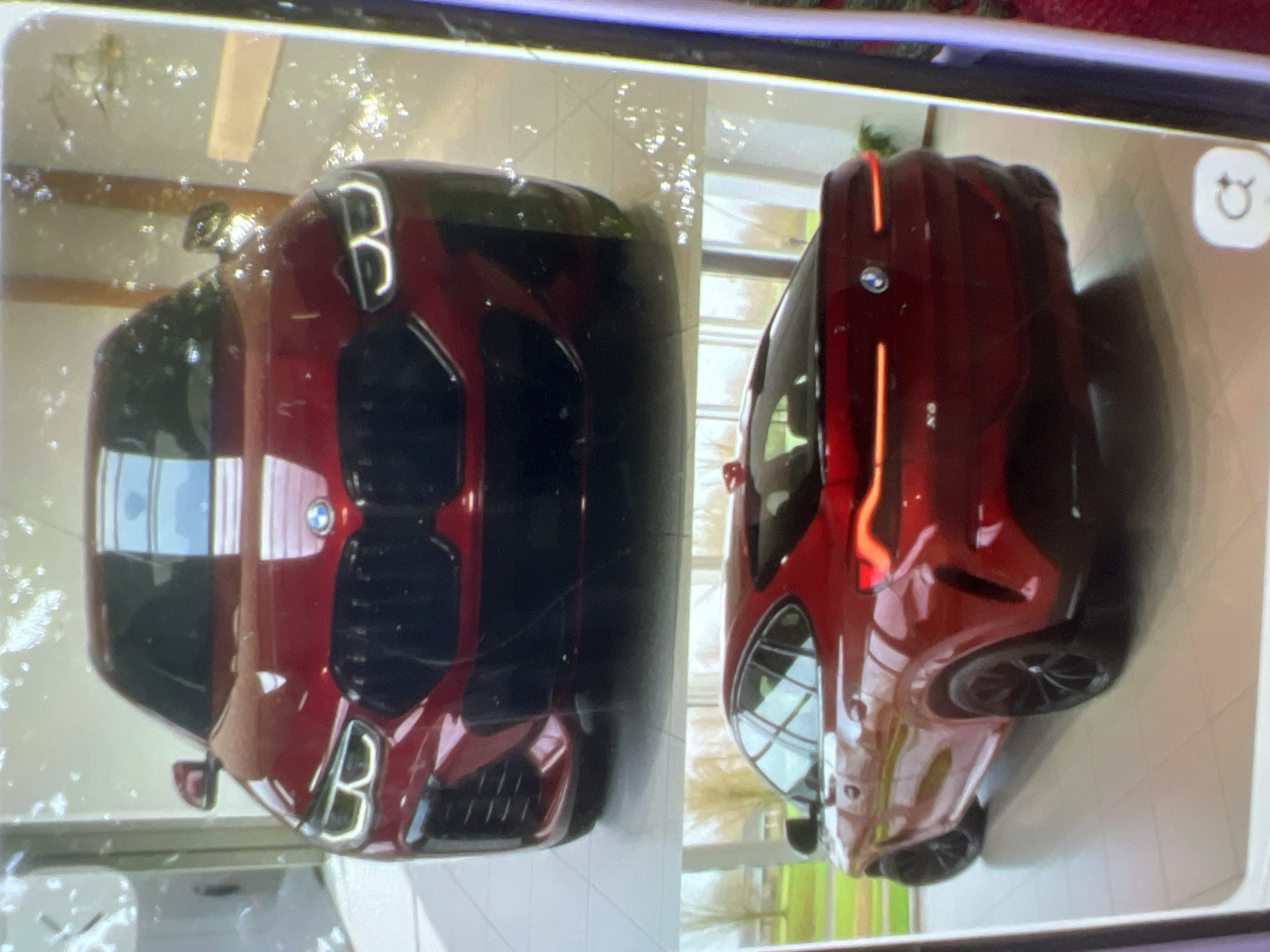 The image displays a red BMW X6 parked indoors, likely in a showroom. The top portion of the image shows the front of the car, highlighting its distinctive grille, headlights, and the BMW logo. The bottom portion shows the rear of the car, emphasizing its taillights, sculpted body, and black wheels. The background of the bottom image includes large windows with a view of trees and what appears to be a residential area, suggesting the showroom is in a suburban or semi-rural setting. There are no people visible in the image. There is no indication of the city of Kara-Dıykan, Kyrgyzstan.