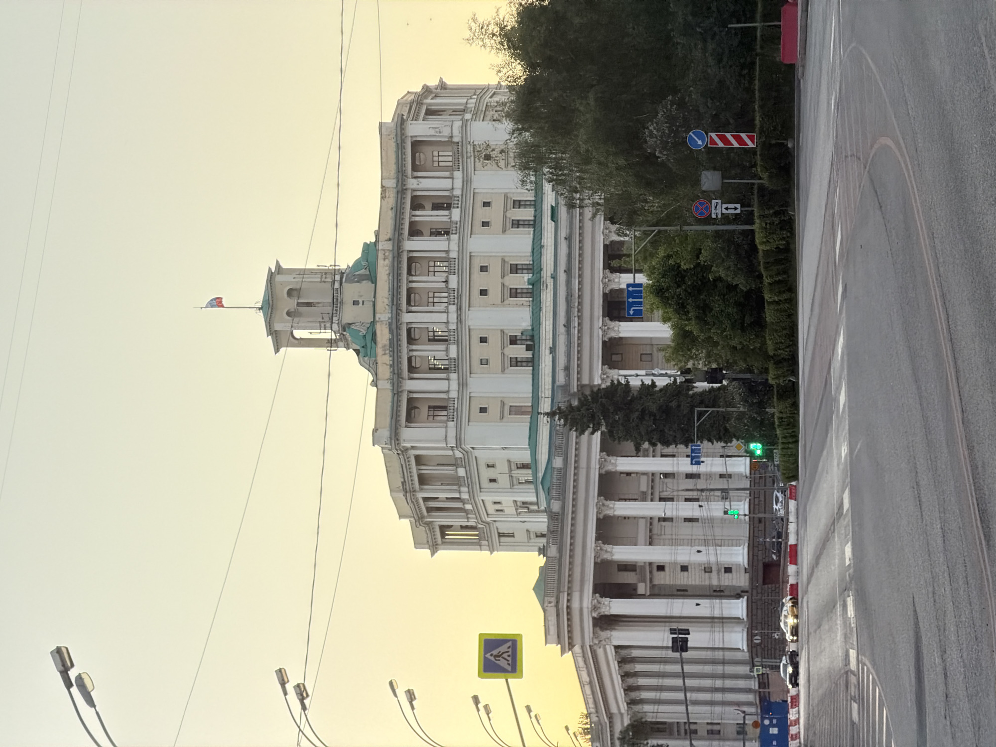 The image captures a daytime scene in Moscow, Russia, focusing on the Mayor of Moscow's office building, a large, multi-story structure with a light-colored facade and a tower with a flag on top. The flag indicates the building is likely a government or official establishment. The building features classical architectural elements like columns and pilasters.
To the right of the building, there is a paved road with typical street markings and several road signs, including a pedestrian crossing sign. There are also trees and greenery present, indicating a street with some landscaping. Streetlights are visible in the lower left. The sky is a pale, slightly yellowish color. There are some vehicles parked along the road.