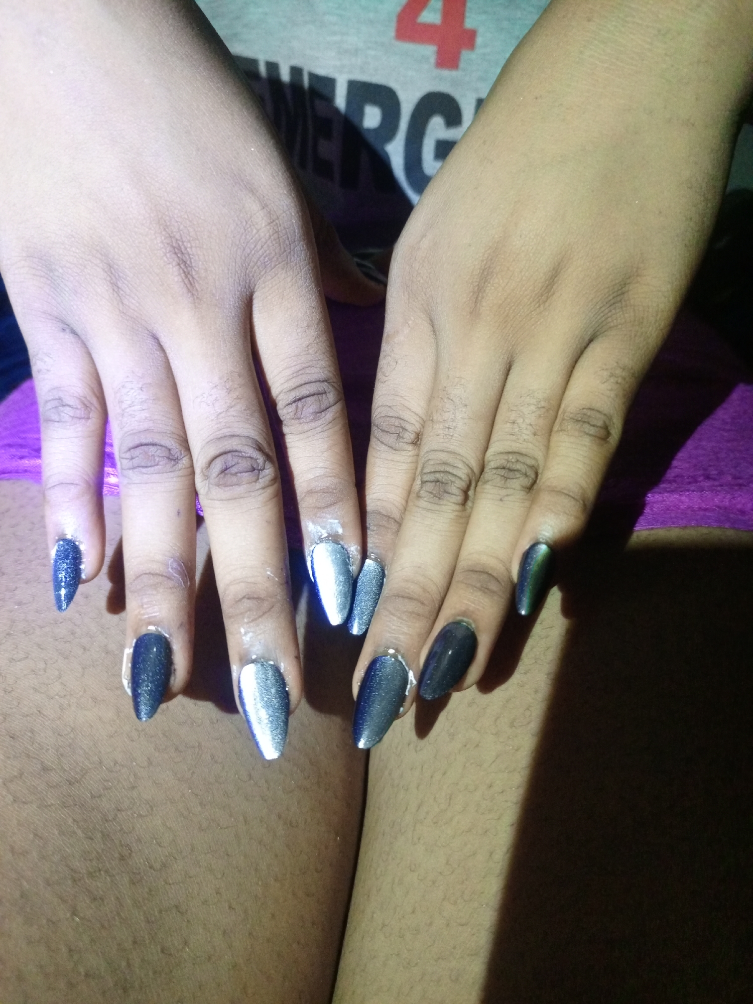 The image shows two hands with long, pointed nails, each painted in alternating shades of dark blue and silver with a glittery finish. The hands are resting on what appears to be a pair of thighs covered in short hair. The background is somewhat obscured, but suggests a partially illuminated environment. The lighting is uneven, casting shadows across the scene. The context of Keffi, Nigeria is not explicitly visible, and cannot be confirmed or denied based on the visual evidence alone.