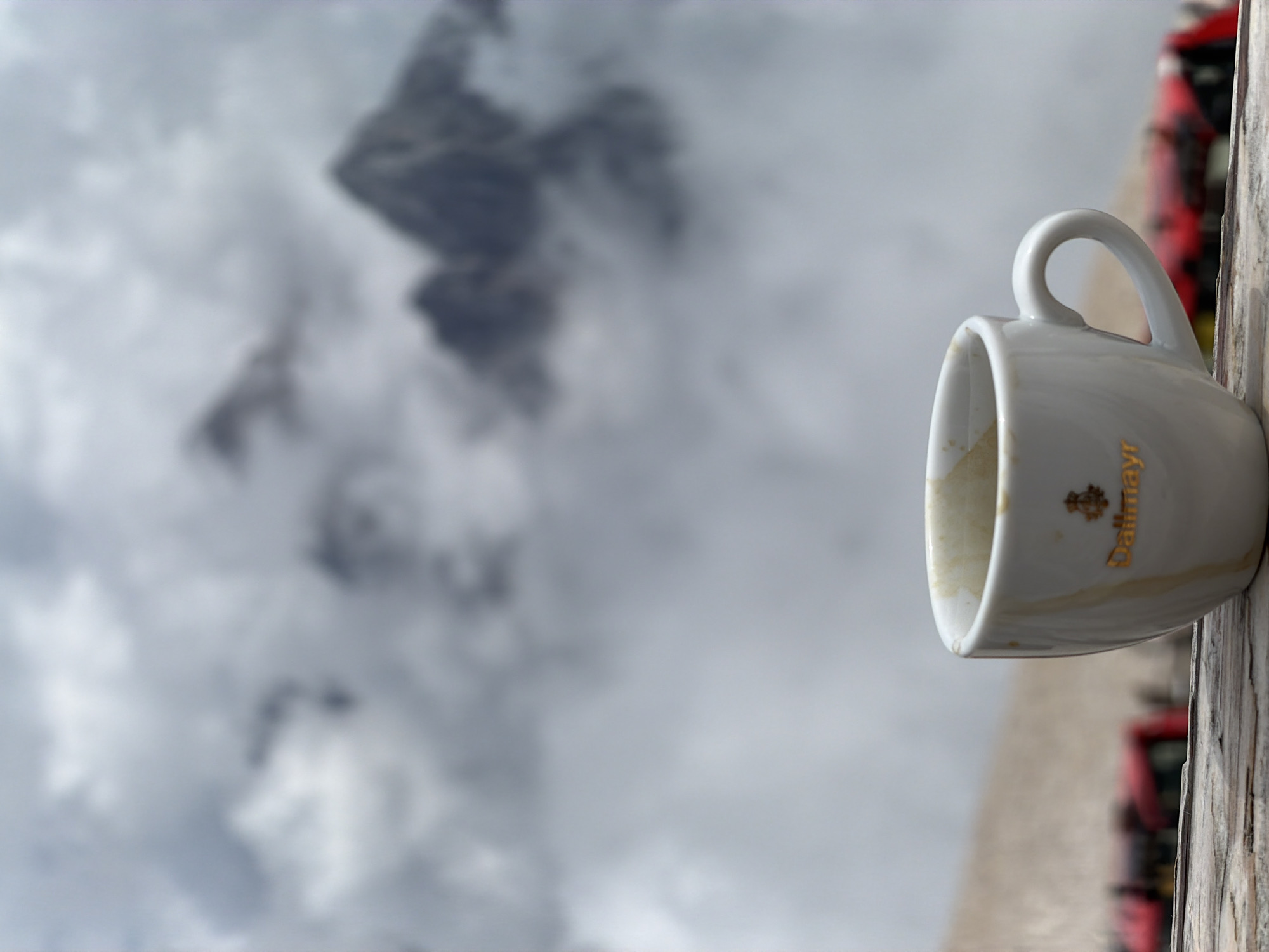 A white coffee cup, with the logo "Dallmayr" visible in gold lettering and an emblem above it, rests on a weathered wooden surface. The cup appears to have been used, with coffee residue staining its interior. In the background, a blurred mountain landscape is visible, shrouded in thick clouds, suggesting a high-altitude location. The muted gray tones of the sky and mountains create a dramatic and atmospheric backdrop. The presence of red objects, also blurred, in the lower portion of the image suggests a possible transportation element, like a train or bus, but they are too indistinct to be identified definitively. The overall scene evokes a sense of peaceful contemplation amidst a majestic natural setting. This image was taken at the Zugspitze, Germany's highest peak, which is accessible from Garmisch-Partenkirchen.