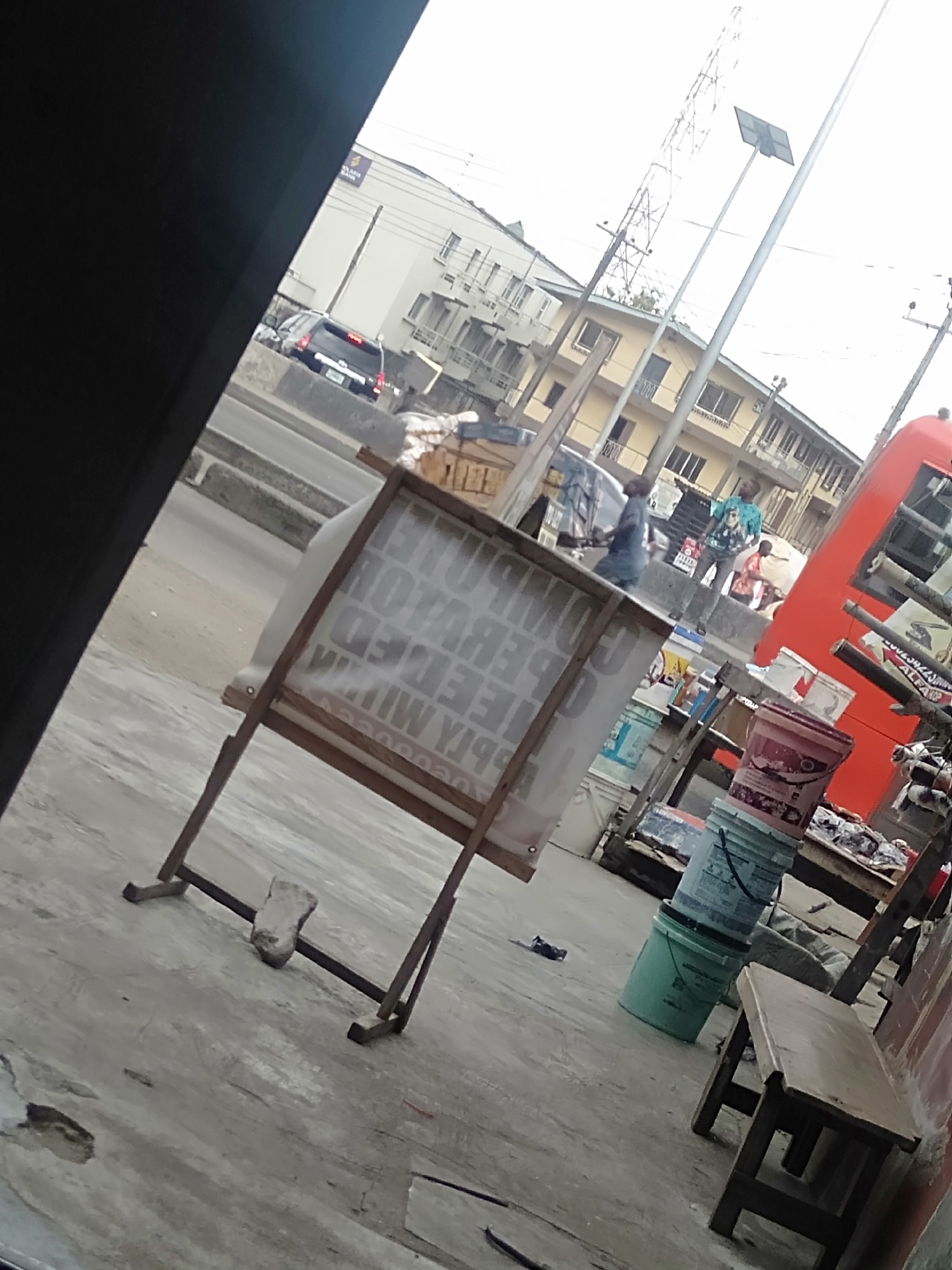 The image depicts a street scene in Shangisha, Nigeria, possibly near a roadside business. Visible elements include:
**Environment:**
- A paved street with vehicular traffic in the background.
- Several buildings in the mid-ground.
- A utility pole with a solar panel fixture.
**Subjects:**
- Several individuals are visible in the background, some appearing to be interacting or walking.
- An individual is located near the roadside business.
**Objects:**
- A wooden-framed sign with a partially obscured advertisement, weighted down with a stone.
- Stacks of plastic buckets.
- A wooden bench.
- A red bus.
**Actions/Interactions:**
- People appear to be walking along the street.
- The roadside business is possibly operating.