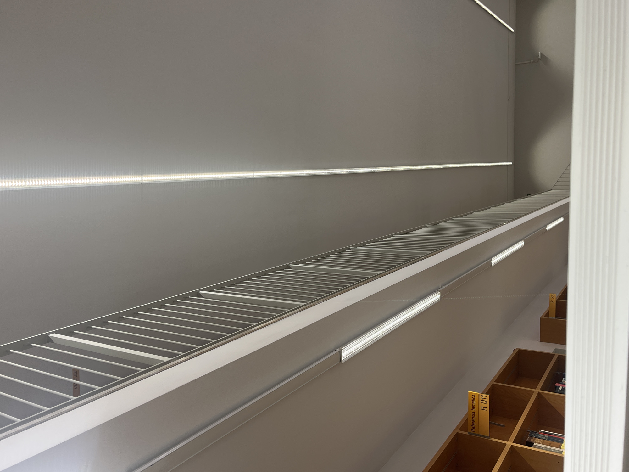 The image captures an upward-looking perspective of a modern, minimalist indoor space, likely a library in València, Spain. The dominant feature is a vast, light gray wall ascending diagonally from the bottom left to the top right of the frame. Long, narrow LED light strips are mounted in linear recesses along this wall and a lower railing, providing bright, even illumination. Some strips clearly show individual LED points, while others present a continuous line of light.
A prominent architectural element, resembling a very long, wide grate or slatted structure made of numerous thin, light-colored bars, runs horizontally across the middle of the frame, also appearing to ascend at an angle parallel to the main wall.
In the bottom right, several light brown, wooden bookshelves with cubby-like compartments are visible, receding into the background along the slanted wall. A tall, white, fluted column or wall section partially obscures the far right. No people or explicit activities are depicted, conveying a sense of quiet order and spaciousness. The bright, diffuse lighting suggests an indoor daytime setting.
Visible text on yellow signs placed within the bookshelves reads: "Referencia temática" and "R 011" on the closer sign, and "R 33" on a sign further back, confirming the library environment.