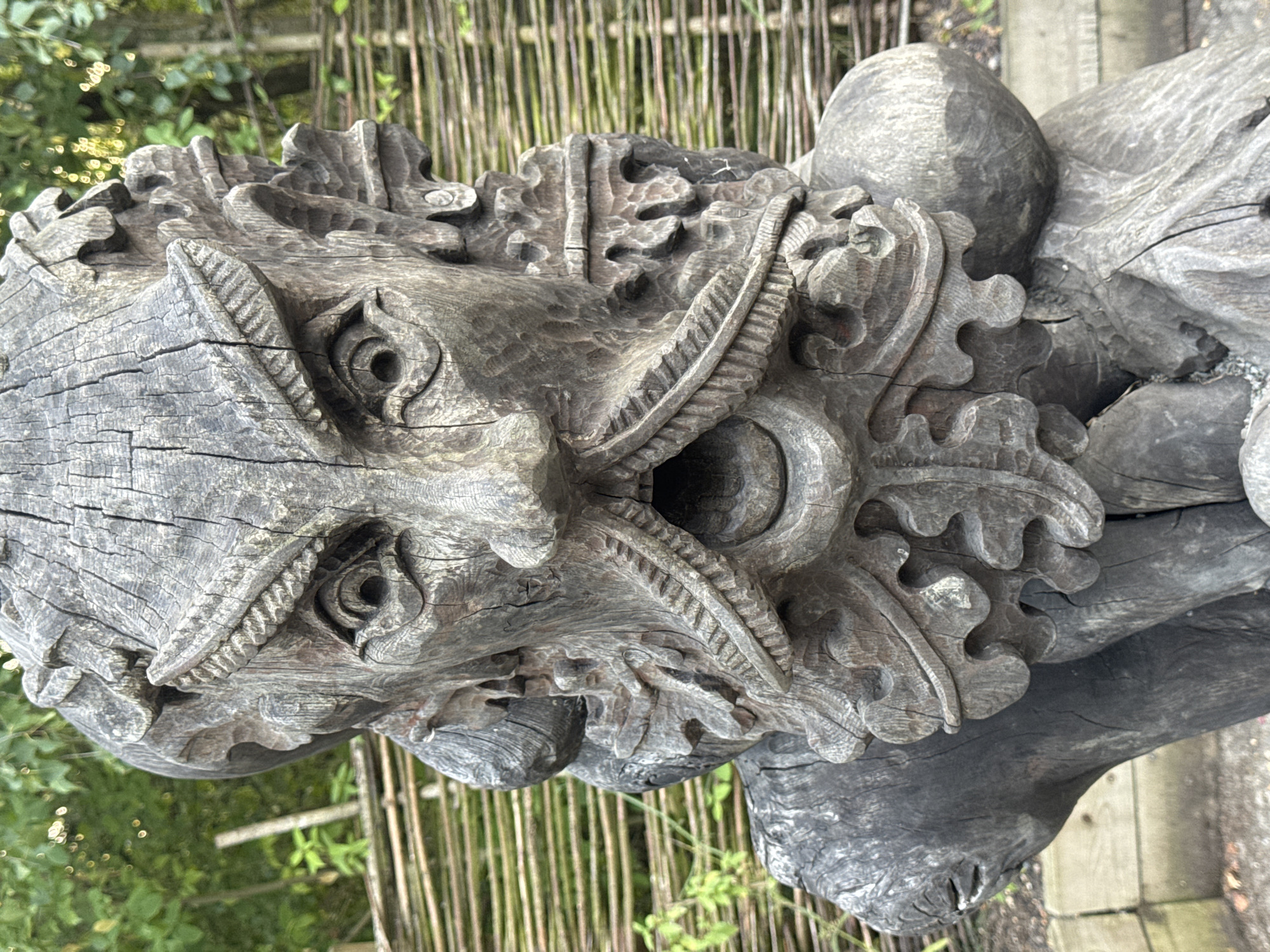 A close-up, eye-level, full shot shows a weathered, grey, wooden sculpture of a face with an open mouth, carved with a grimace. The sculpture features a prominent nose, furrowed brow with carved eyebrow ridges, and a detailed mustache and beard. The eyes are deeply set within the carved sockets, and there are carved ears on either side of the head. The wood is cracked and aged, showing the grain and texture of the material. The background consists of a blurred, green leafy foliage and a light brown woven fence. The sculpture appears to be outdoors. This specific sculpture is located in the city of Gravesham, United Kingdom.