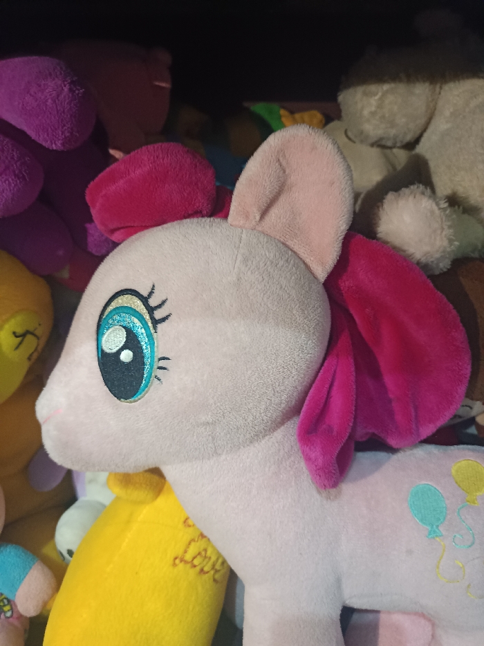 The image displays a close-up, left-profile view of a light pink plush pony toy. The toy features an embroidered left eye with a black pupil, a teal iris with white highlights, and black eyelashes. A faint pink embroidered nostril is visible on the muzzle. A darker pink fabric component forms the mane, positioned on the top of the head and behind the light pink inner ear. The toy's body is made of a soft, short-pile fabric. On the visible flank, an embroidered 'cutie mark' depicts three balloons: one teal and two yellow, with yellow strings trailing downwards. The pony is situated within a densely packed assortment of other plush toys. Visible in the background are a purple plush toy on the upper left, a bright yellow plush toy with brown embroidery on the lower left, and various indistinct beige and darker-colored plush items. The scene is observed under ambient indoor lighting. This location is in Gotha, Germany.