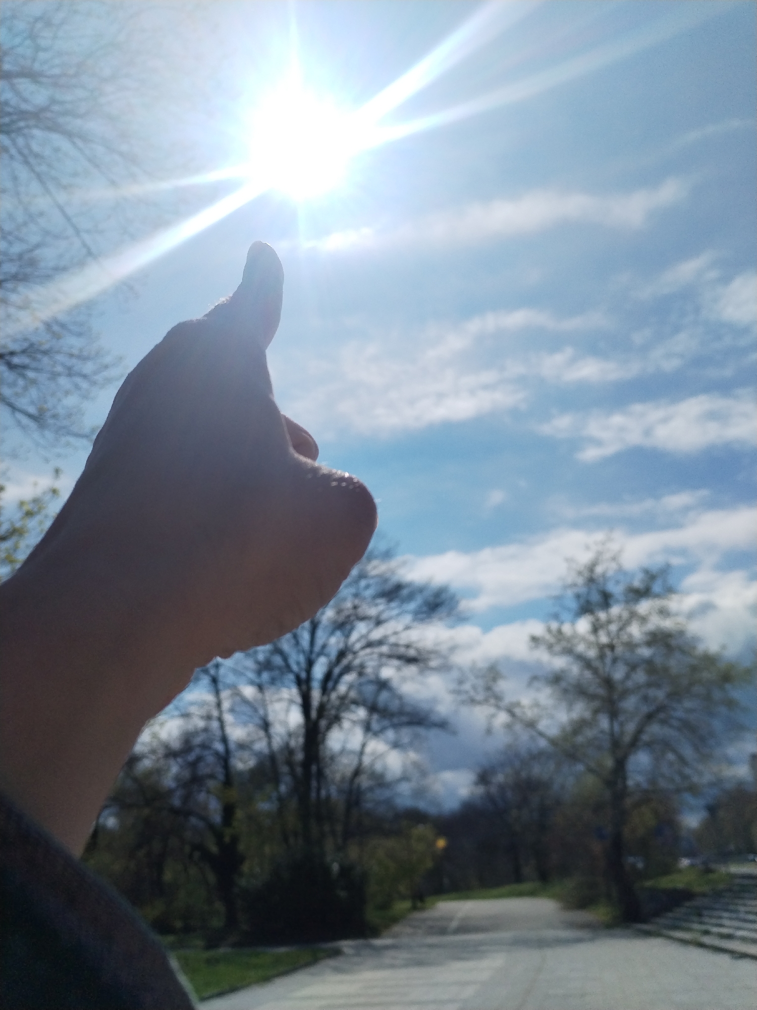 The image displays a human hand in silhouette, making a "thumbs up" gesture towards a bright sun positioned in the upper center of the frame. The sun, a powerful light source, creates a strong lens flare and glare across the upper portion of the clear blue sky, which also features scattered white clouds. The hand is illuminated from behind, highlighting its outline against the intense light.
The background reveals an outdoor setting, likely a park or natural area. Numerous trees, some with bare branches and others showing nascent green foliage, suggest a springtime scene. A paved path or road curves through the lower part of the image, leading into the distance. On the far right, a section of what appears to be concrete stairs is visible, further defining the landscape.
The overall impression is of a sunny day, with the time of day likely morning or late afternoon given the sun's angle and intensity. The "thumbs up" gesture universally conveys approval, positivity, or well-being. No discernible text is visible within the image. The setting is contextually located in Varna, Bulgaria.
