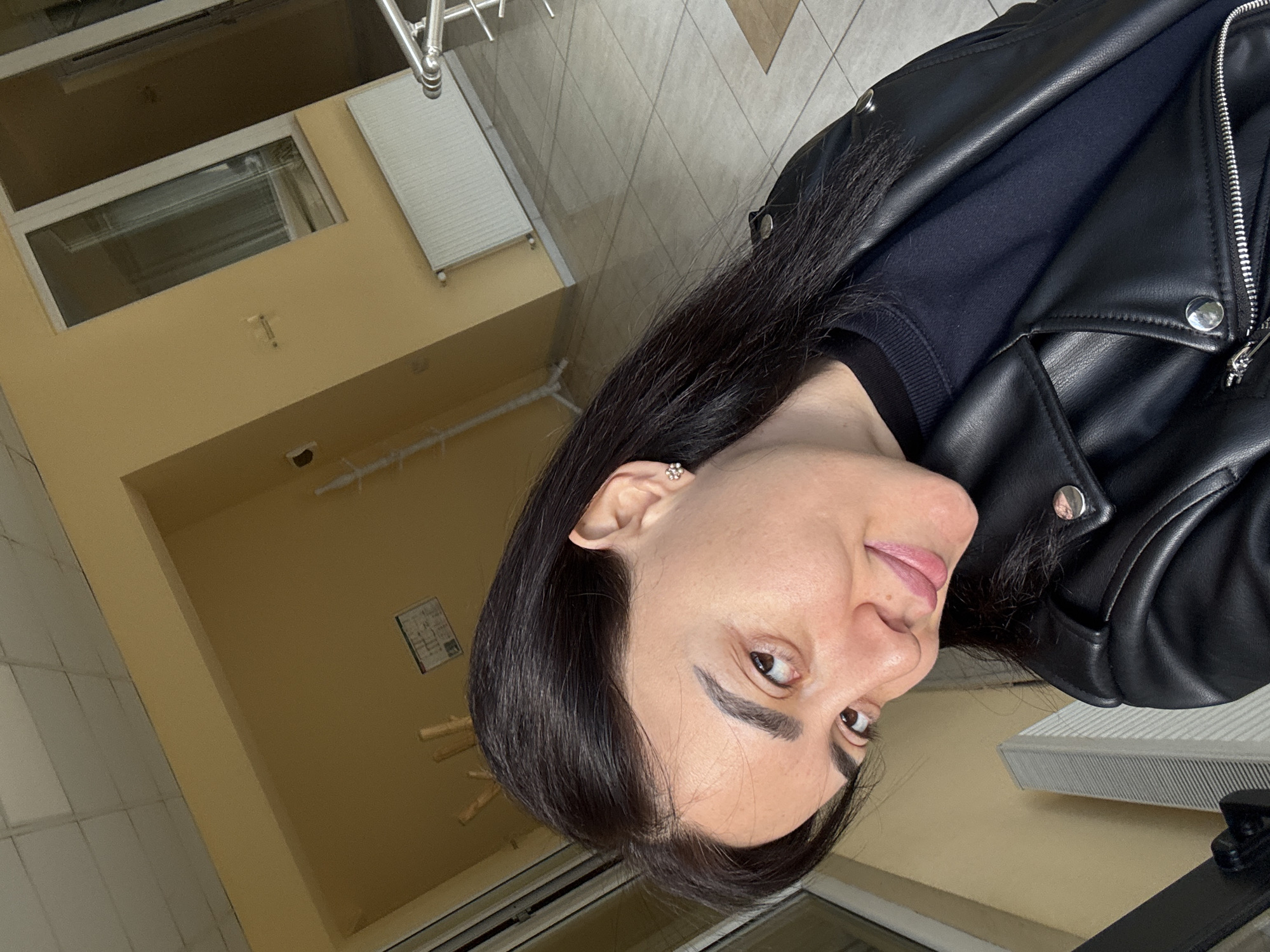 A woman with dark hair and fair skin is wearing a black leather jacket and a dark shirt. She is looking directly at the camera with a slight smile. She is wearing small flower-shaped earrings. The background is a room with yellow walls and tiled floors. There is a window and a radiator on the wall. A piece of furniture is in the foreground. The photo appears to be taken indoors. There is no explicit indication of the city of Tbilisi, Georgia.