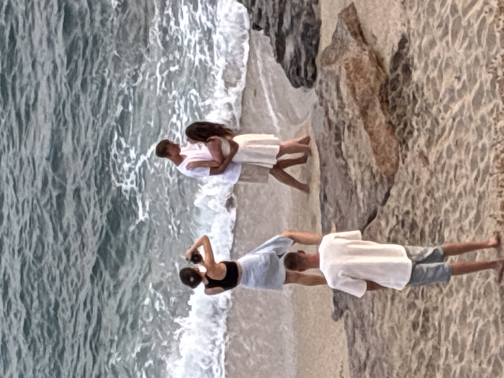 A photograph depicts a group of people on a sandy beach with the ocean in the background. The image is rendered in a painterly style, with soft edges and muted colors.
In the foreground, a person wearing a white t-shirt and denim shorts stands facing away from the camera, looking towards the ocean.
In the mid-ground, a couple is embracing near the water's edge. The man is wearing a white t-shirt and light-colored shorts, and the woman is wearing a light-colored dress. They are positioned in a romantic pose. To their left, a woman wearing a black tank top and a light-colored skirt is holding a camera and appears to be taking a photo.
The background consists of the blue-grey ocean with waves breaking on the shore. Rocky formations are visible on the right side of the image. The overall scene suggests a casual outing at the beach.
The image does not contain any elements that would specifically identify the location as Alanya, Turkey.