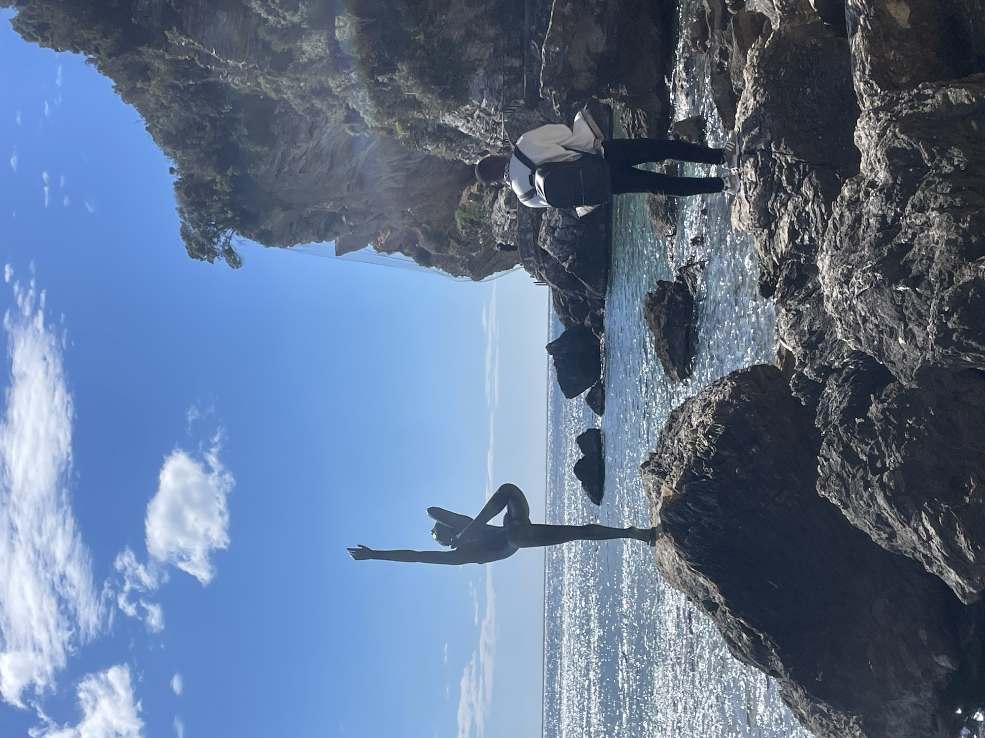 The scene depicts a coastal landscape in Budva, Montenegro, specifically near the Old Town, featuring the "Dancer of Budva" statue. In the foreground, a dark bronze statue of a female dancer in a ballet pose, with one leg bent and one arm extended upwards, stands on a large, dark rock at the water's edge, facing the open sea.
To the right and slightly behind the statue, an individual is observed from the rear, standing on a jagged dark rock. This person wears a light-colored, long-sleeved top, dark trousers, light-colored athletic shoes with dark stripes, and a dark backpack. They are looking towards the sea or the adjacent cliff.
The sea extends into the background, characterized by intense sun glare reflecting off its surface and dark blue-green water near the shore. Numerous dark, irregular rocks are scattered along the coastline and protrude from the water. A large, steep, rocky cliff face, topped with dense green vegetation, dominates the right side of the image. A safety net is visible along a section of the cliff, and a narrow path with small, indistinct figures can be seen on its lower part. The sky is clear blue with scattered white cumulus clouds, indicating bright daylight.
