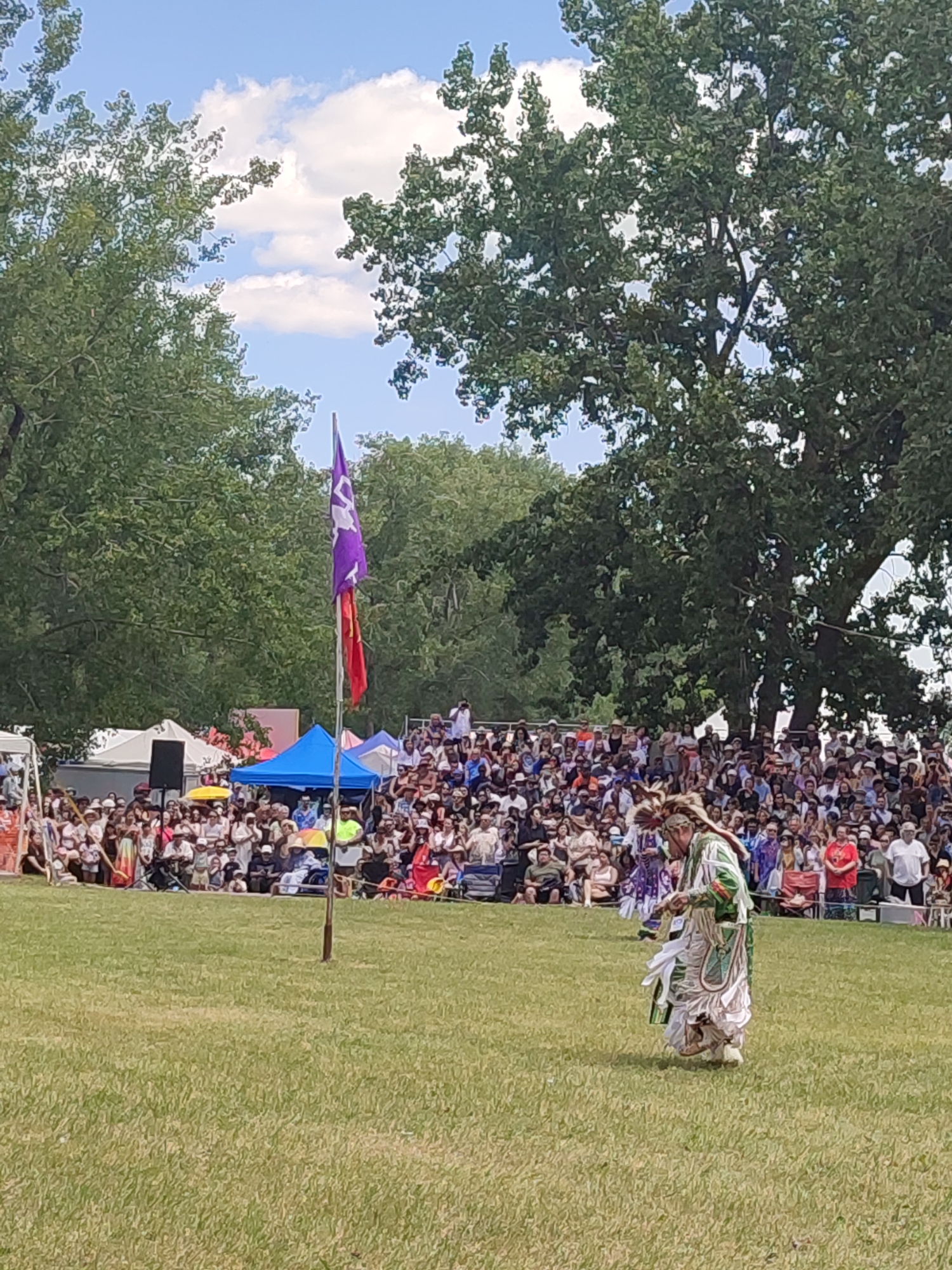 The image shows an outdoor gathering in Kahnawake, Canada, with a large crowd of spectators seated on bleachers and on the grass. In the foreground, a person in traditional Indigenous regalia, adorned with feathers and intricate beadwork, is performing a dance. The person is wearing a long, flowing skirt and a decorated top. In the background, two flags are visible on a pole, one purple with a white emblem and another red. Tents and structures suggest a festival or event setting. Lush green trees frame the scene under a partly cloudy blue sky.