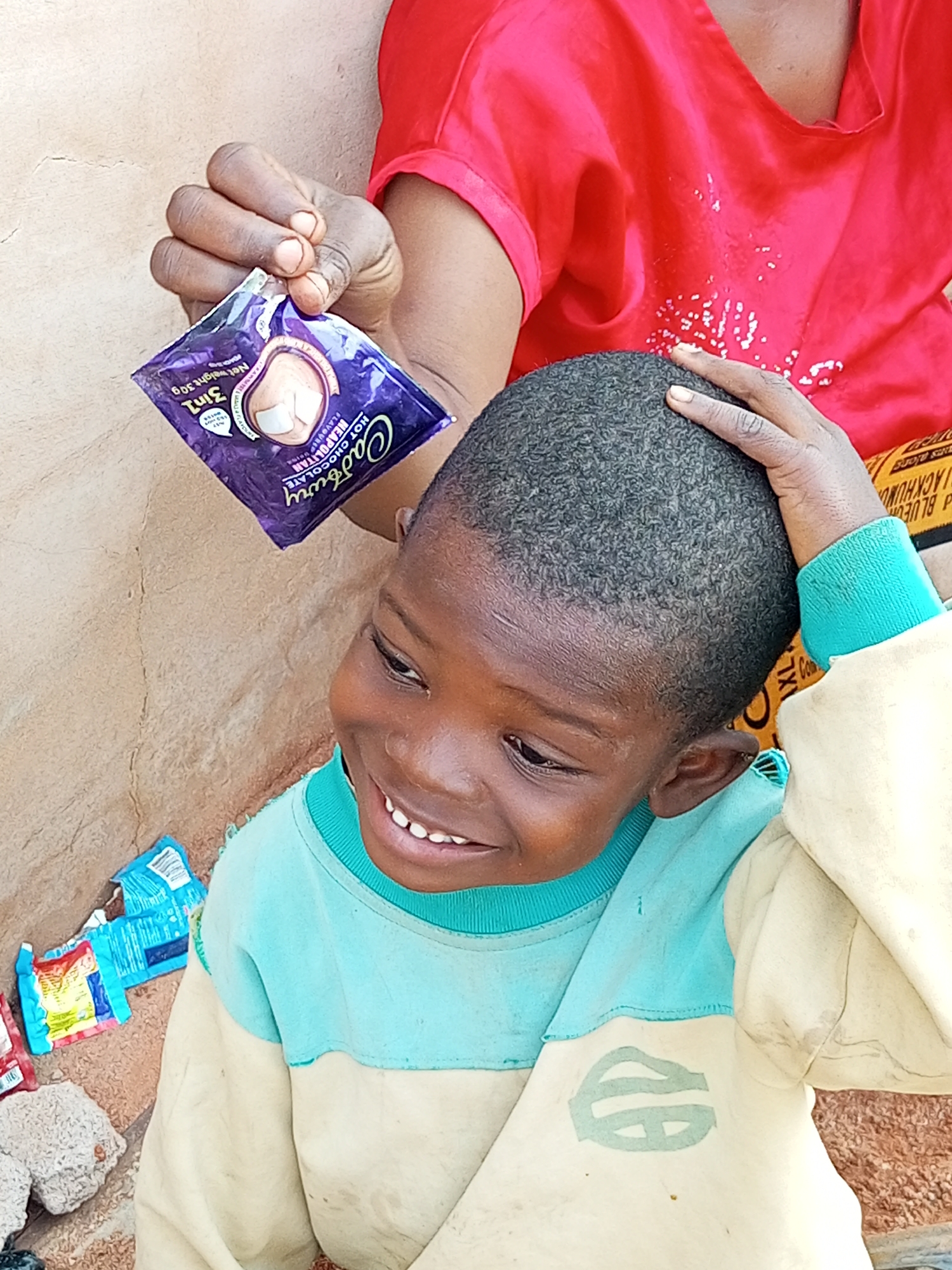 In Tempure, Nigeria, a young child with dark skin and short, dark hair is being held by an adult wearing a red top. The child is smiling and has one hand resting on their head. The adult is holding a small purple packet of Cadbury hot chocolate in their hand. The background is a textured wall. There are also some discarded packets on the ground near the child. The scene appears to be outdoors, as indicated by the lighting and the outdoor setting.