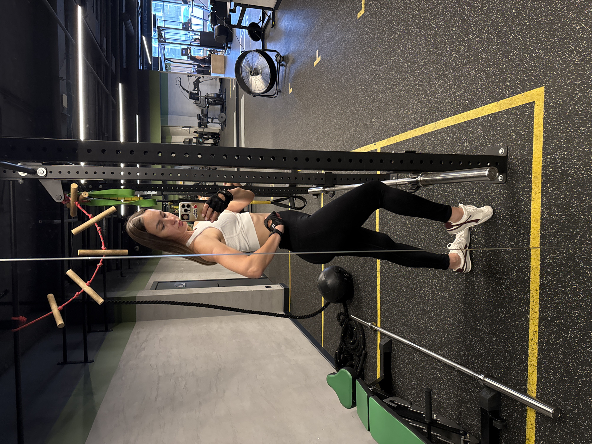 A woman is lying on a gym floor, performing an exercise. She is wearing a white sports bra and black leggings, with black fingerless gloves on her hands and white athletic shoes on her feet. Her body is positioned horizontally, with her head to the left and feet to the right. She is looking at her phone, which she is holding in her right hand. The phone case has a sticker with hearts and text that says "Team".
The setting is a modern gym with a black rubberized floor marked with yellow lines. A black metal squat rack is in the foreground, with the woman positioned beneath it. To the left of the squat rack, there are wooden rungs attached to red ropes, part of a climbing apparatus. In the background, various gym equipment is visible, including weight machines and a large industrial fan. The lighting in the gym appears to be artificial, with bright fluorescent lights.
The woman appears focused on her phone, with a relaxed expression. The gym seems to be well-equipped and maintained. There are no clear indicators of the time of day or weather. The location is identified as Sochi, Russia.