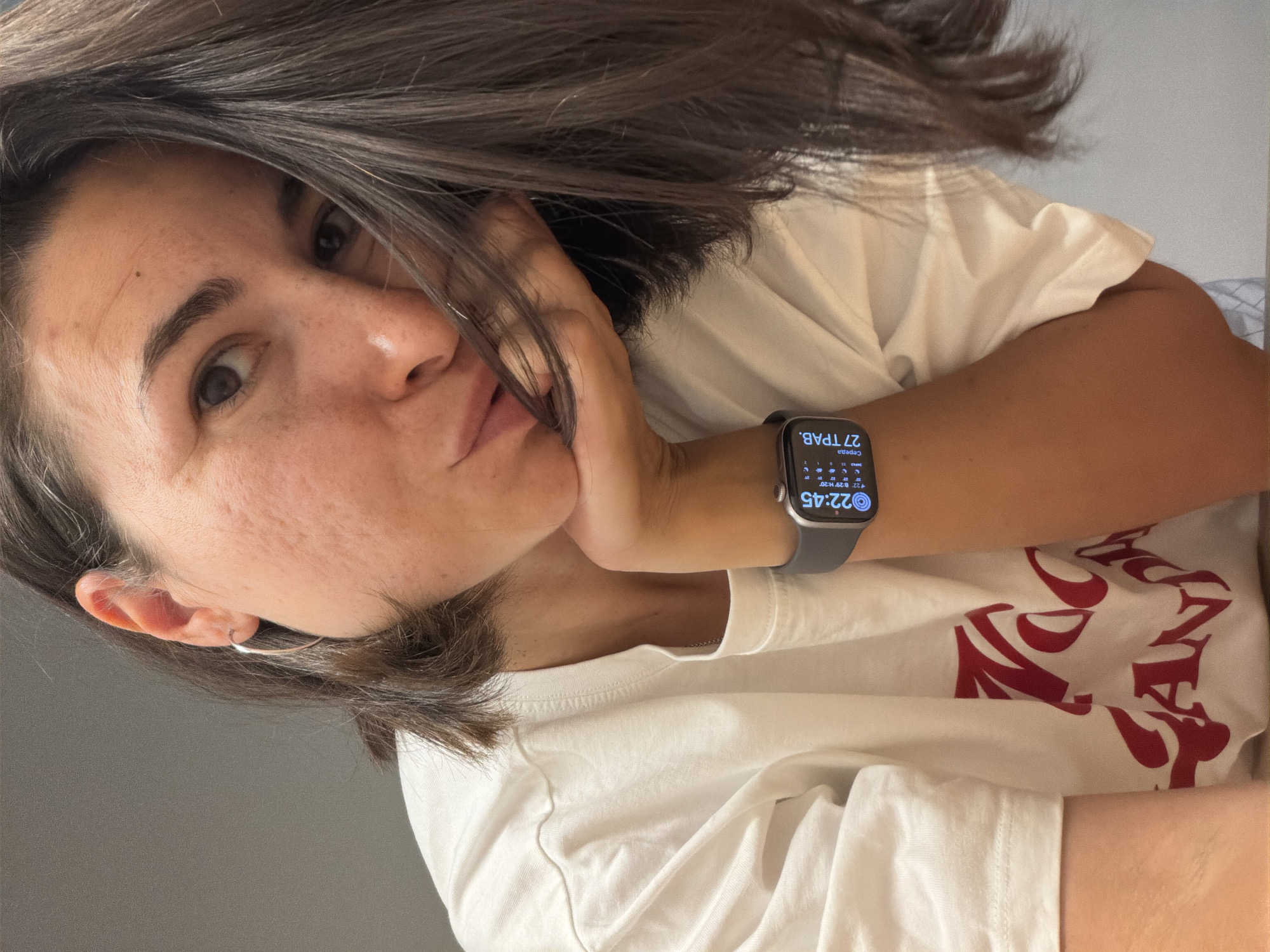 A woman with dark hair is looking at the camera, resting her face on her hand. She is wearing a white t-shirt with a red design. An Apple Watch is visible on her wrist, displaying the time 22:45 and the date 27. Her location is unknown.