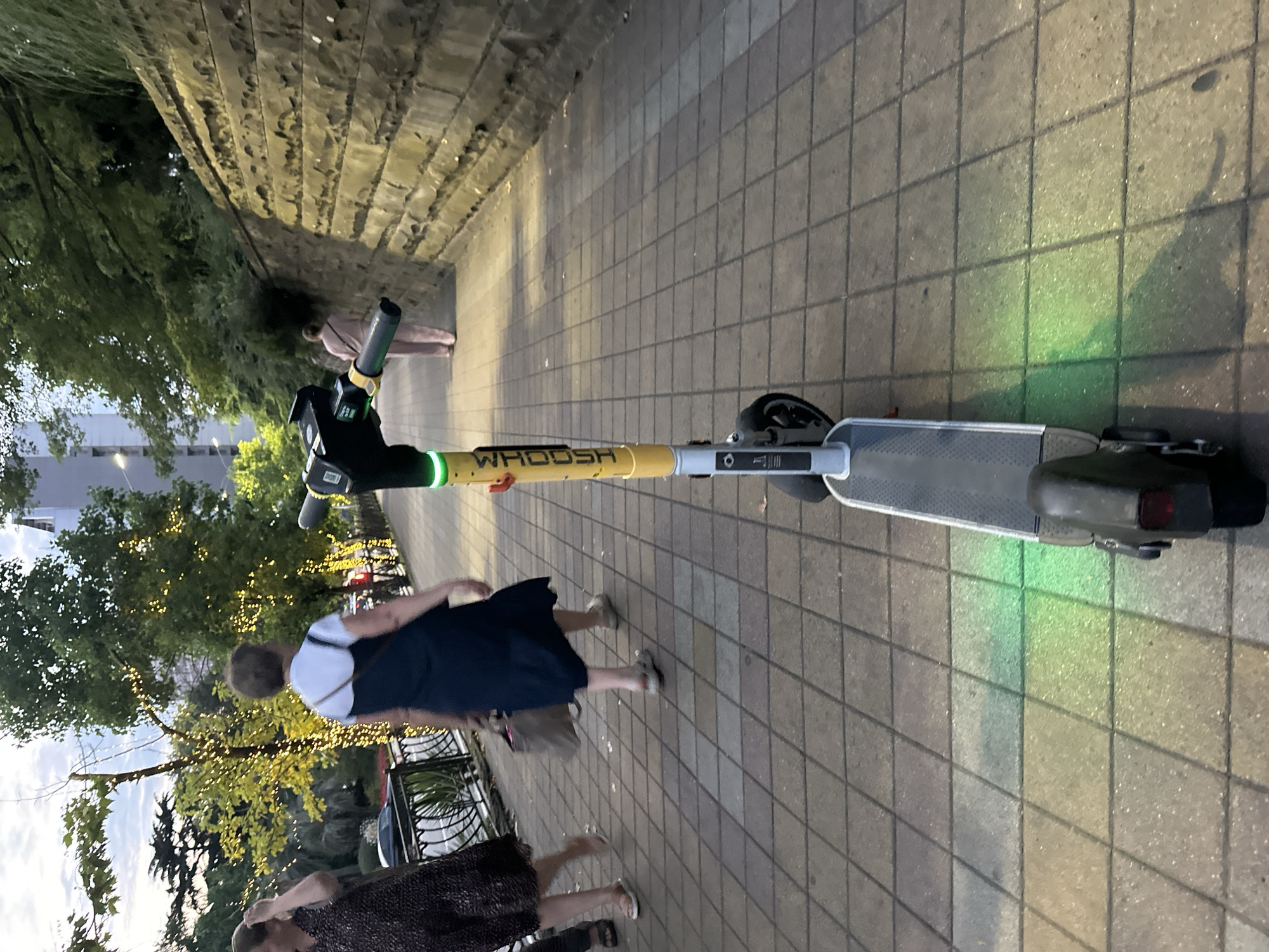 In the city of Unknown, Unknown, an electric scooter with "WHOOSH" branding is parked on a tiled sidewalk. The scooter has a yellow pole with black handlebars and a green illuminated ring. Its front wheel is visible, and its rear wheel is obscured. Green light is casting a shadow on the sidewalk from the scooter's base. In the background, a woman in a dark patterned dress walks away from the camera, and another woman in a dark blue dress with a white strap is walking in the same direction. Further back, a person in light pink clothing is walking away. The sidewalk is bordered by a stone wall on the right and trees with decorative lights on the left. The scene appears to be outdoors, possibly in the evening, with the sky visible in the upper left corner.