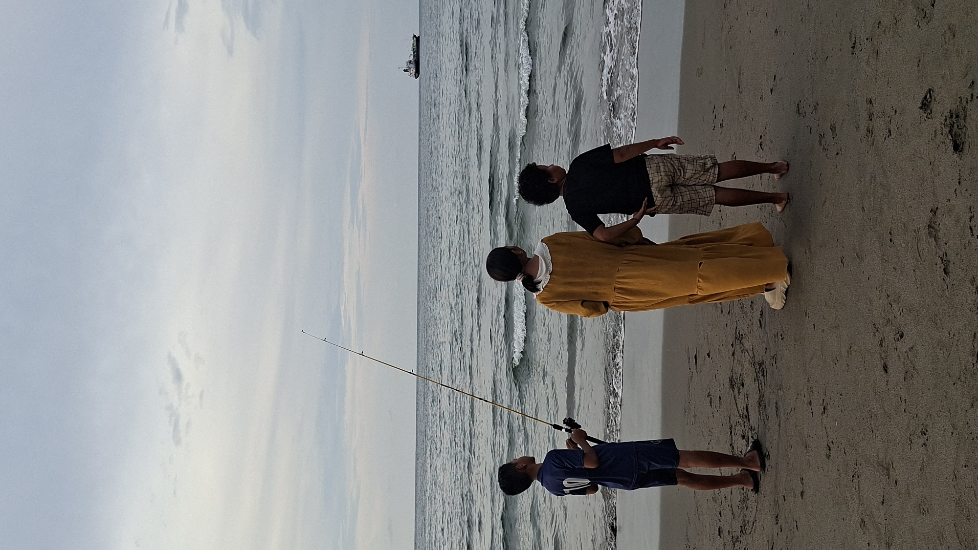The image depicts three children on a sandy beach in Lhokseumawe, Indonesia. The children are standing facing the ocean, with their backs to the camera. The youngest, a boy, is on the left, wearing a blue t-shirt with the number 10 on it and dark shorts, holding a fishing rod. In the center is a girl in a long, mustard-yellow dress with a white collar. To the right is another boy, wearing a black t-shirt and checkered shorts. The sea is calm with gentle waves breaking on the shore. In the distance, a cargo ship is visible on the horizon under a cloudy sky. The sand appears damp near the water's edge and drier further back. The overall impression is one of a tranquil, end-of-day scene.