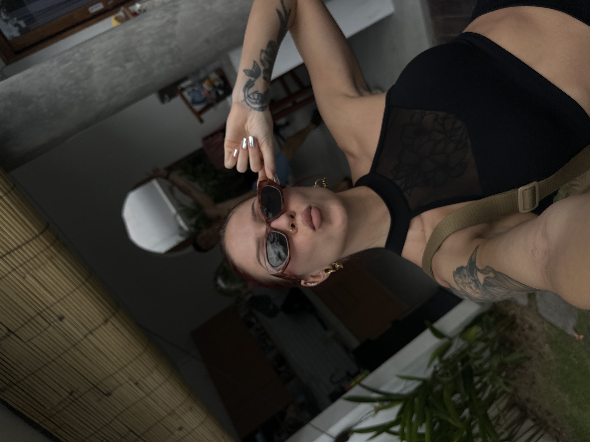A selfie from North Kuta, Indonesia, shows a woman in a black top and sunglasses. She is indoors near a bamboo-like screen, with glimpses of greenery visible. She has visible tattoos on her arms and gold earrings.