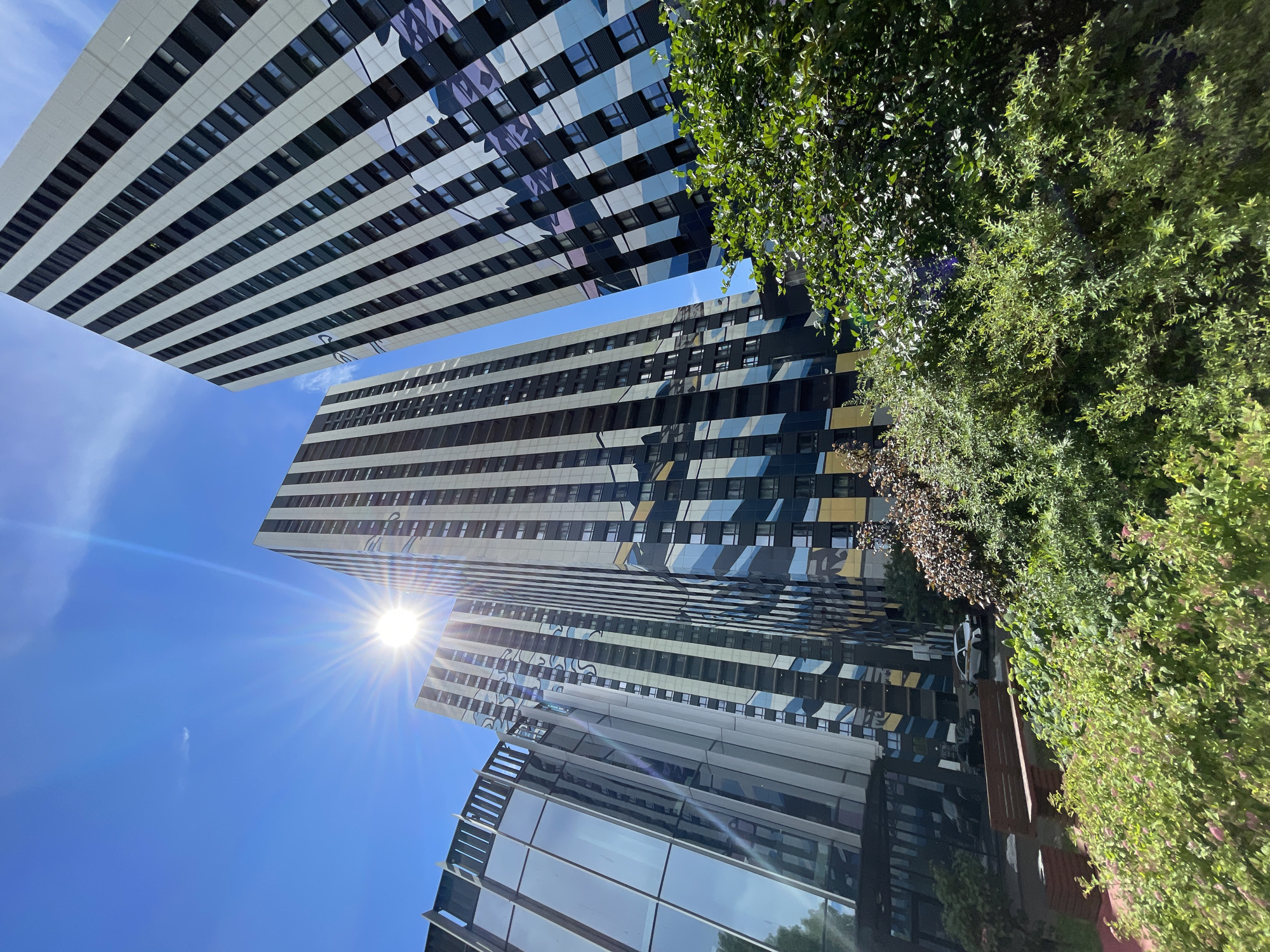A low-angle shot captures three modern, tall buildings with dark and white striped facades, reflecting the bright sun and blue sky. The buildings have large windows and geometric patterns in blues, purples, and yellows. In the foreground, lush green bushes and trees partially obscure the lower portions of the buildings and a paved area with a wooden bench and two dark-colored trash bins. A white car with orange and black accents is parked in the background, along with a dark-colored car. The scene is likely a residential or commercial complex in Moscow, Russia, given the architectural style.