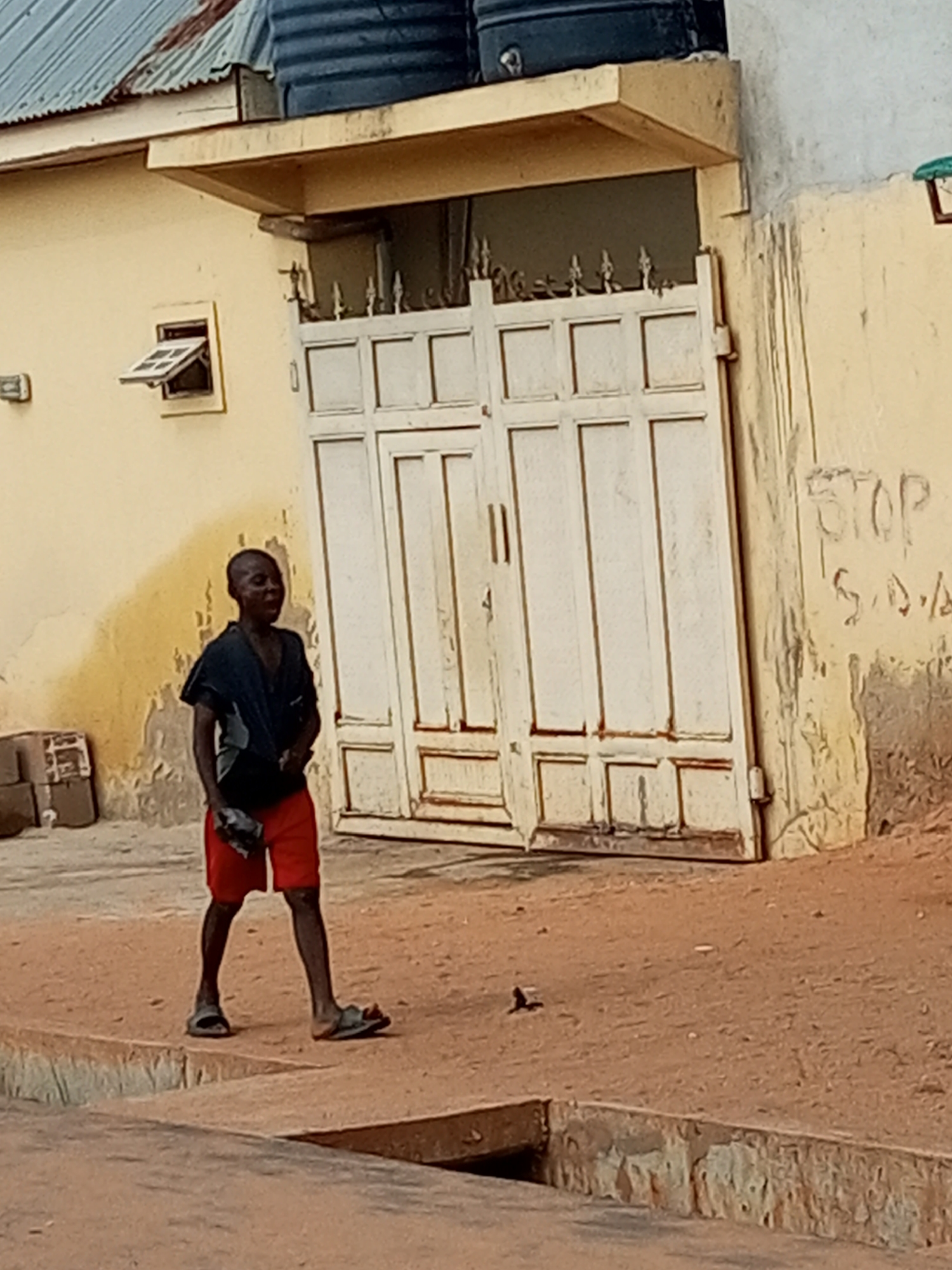 A young boy is captured in a candid moment, walking on a dirt path in front of a building. He appears to be in his early teens, with dark skin and short, dark hair. He is wearing a dark, loose-fitting t-shirt and bright red shorts, along with dark sandals. In his left hand, he clutches a dark object, possibly a piece of cloth or a small bag. His facial expression is one of slight amusement or surprise, with his mouth open.
The background reveals a weathered, yellow building with a corrugated metal roof. A large white double gate dominates the center of the scene, showing signs of age with peeling paint. Above the gate, there's an overhang and two large black water tanks. To the left of the gate, a small window with a louvre shutter is open. Graffiti, including the word "STOP," is visible on the wall to the right of the gate. The ground is a reddish-brown dirt, with some scattered debris and a drainage channel in the foreground. The overall setting suggests a residential area in Tempure, Nigeria.