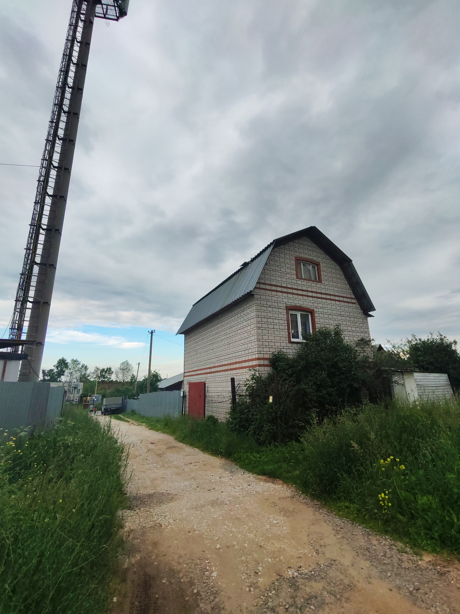In the city of Pervomaysky, Russia, a gravel road winds through a sparsely populated area under a cloudy sky. To the left, a tall, industrial tower with a ladder dominates the foreground, its metal structure contrasting with the natural surroundings. Adjacent to the tower, a grey metal fence marks the boundary of a property. A two-story house constructed from light-colored bricks with red horizontal accents stands on the right side of the road. This house features a dark metal gabled roof and two windows visible on its facade, one on the upper floor and another on the lower. In front of the house, dense green bushes and overgrown vegetation line the property. Further down the road, more houses and fences can be seen, along with a truck and a few individuals walking. The ground is covered with grass and wildflowers, suggesting a rural or semi-rural setting.