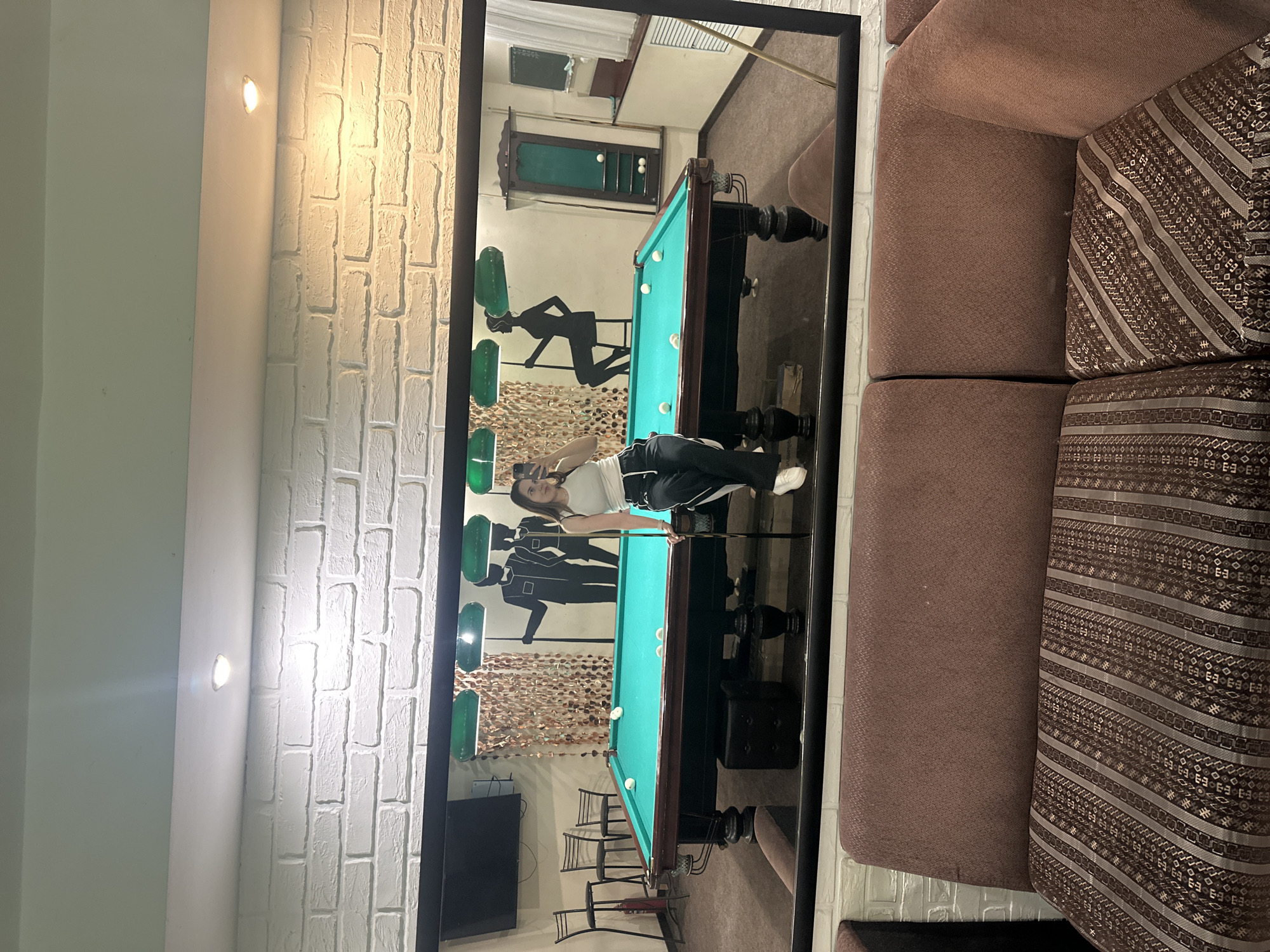 The image shows a young woman taking a selfie in a mirror in a billiards hall. She is wearing a white tank top and black athletic pants with a white stripe. She is leaning against the edge of a billiards table and holding a phone in her right hand. The billiards table is green with white balls. In the background, there are silhouette graphics of people playing billiards on the wall, as well as several green lamps hanging from the ceiling. A TV screen is visible on the wall to the left of the table, and several chairs are arranged nearby. The room appears to be decorated with white brick wallpaper and has a brown couch in the foreground. The location is identified as a billiards hall in the city of Perm, Russia.