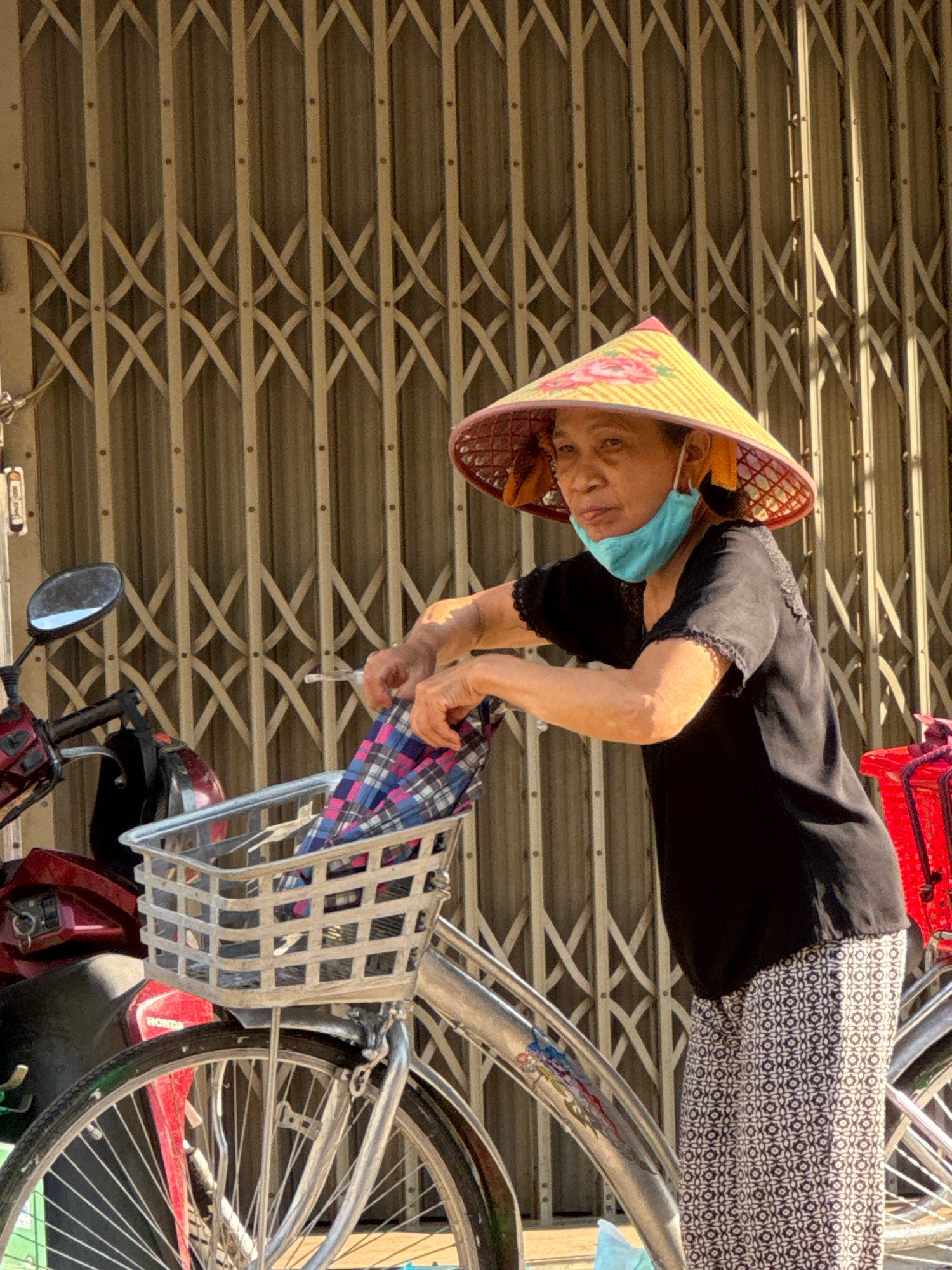 A Vietnamese woman wearing a conical hat and a light blue face mask is standing next to a bicycle, in front of a corrugated metal gate. She is wearing a black short-sleeved shirt and patterned pants, and is reaching into the bicycle's basket, which contains a plaid cloth. A portion of a red motorcycle is visible to the left of the bicycle, and a red basket is attached to the rear of another bicycle to the right. The scene appears to be a street in Nha Trang, Vietnam, given the commonality of bicycles, motorcycles, and the style of the gate.