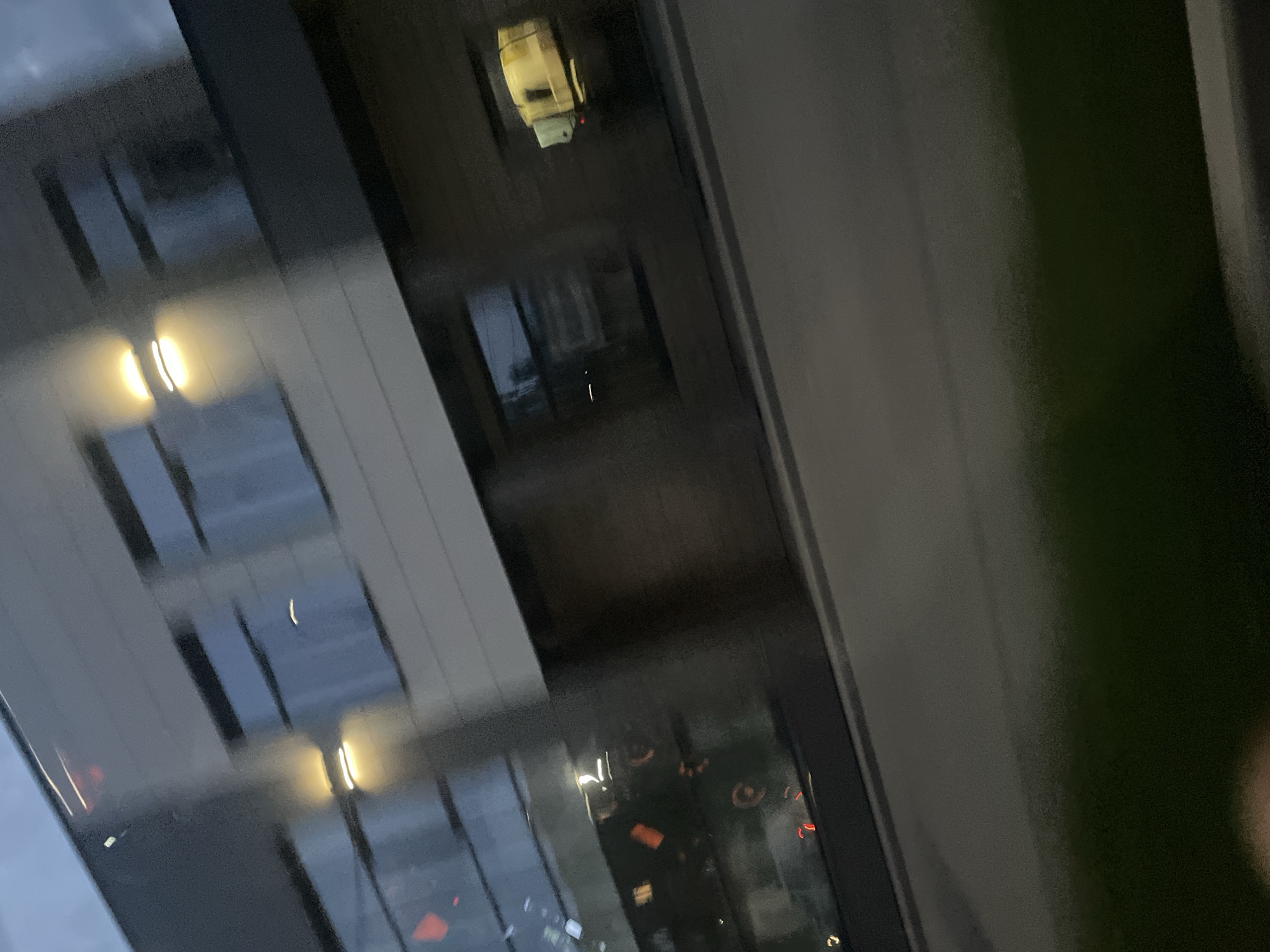 The image shows a blurred exterior view of a modern building with large windows, possibly in Votkinsk, Russia. The building appears to have multiple stories, with some windows lit from within. There is a dark horizontal line across the lower portion of the image, suggesting a road or sidewalk. The sky is overcast and appears to be dusk or evening. The overall impression is one of movement, likely captured from a vehicle passing by.