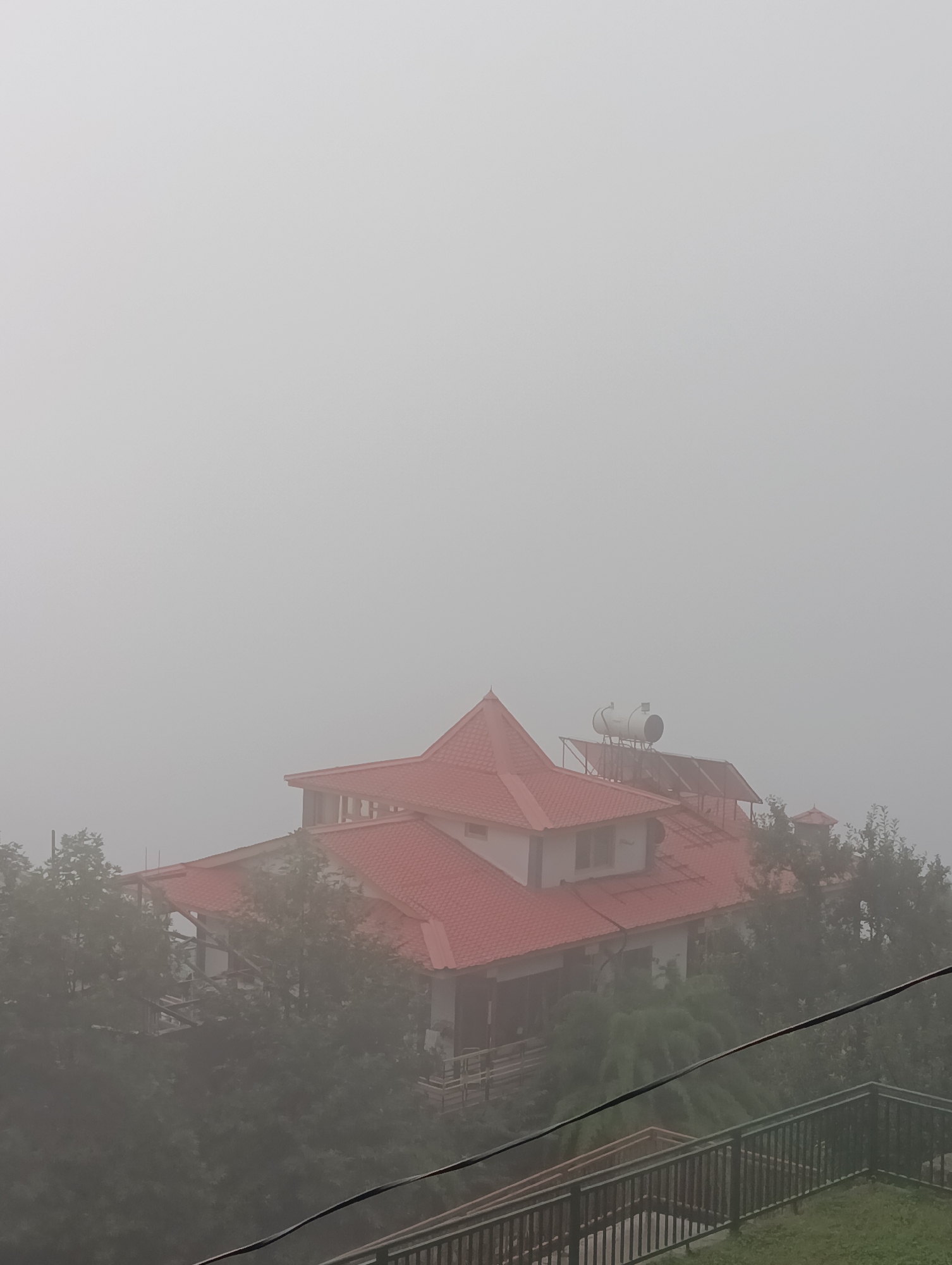 The image shows a building with a red tiled roof, partially obscured by fog. The building appears to be situated on a hillside, with trees visible in the foreground and surrounding the structure. A solar water heater is mounted on the roof. The sky is overcast and a dark cable runs diagonally across the lower right portion of the image, with a metal railing and stairs below it. The scene is set in the city of Chhyal, India.