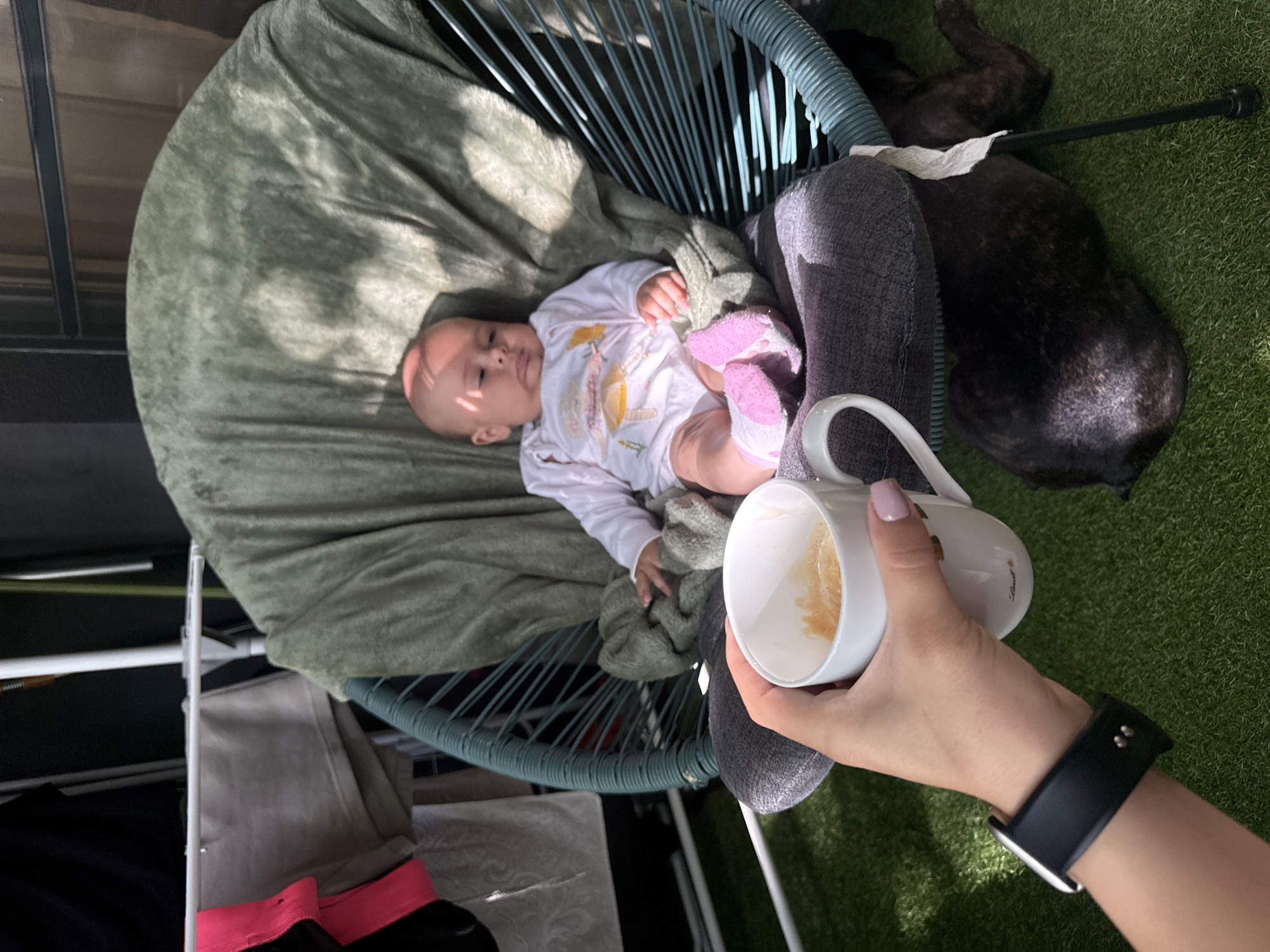 A baby is sitting in a green chair covered with a blanket. A person's hand is holding a cup of coffee in the foreground. A dog is lying on the ground next to the chair. The scene appears to be on a balcony or patio with artificial grass. There are clothes drying on a rack in the background.